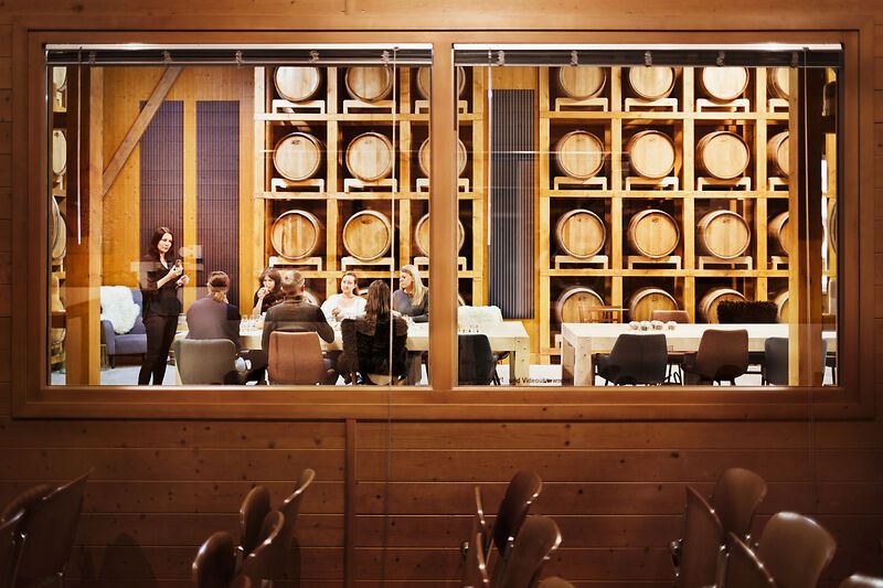 Blick durch ein Fenster in einen Raum mit Whiskyfässern und einer Gruppe von Menschen, die an einem Tisch sitzen.