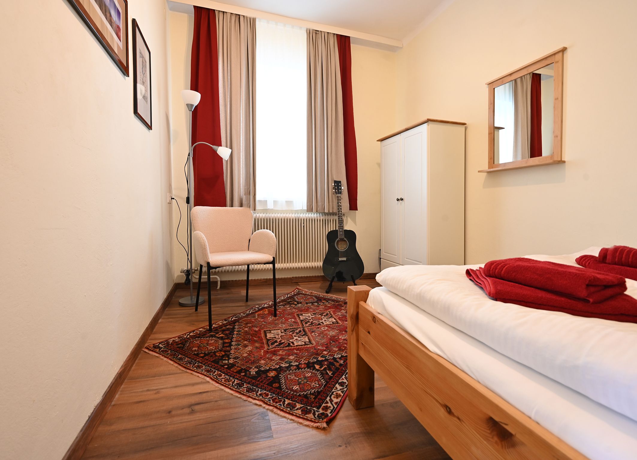 Kleines Zimmer mit Bett, Teppich, Stuhl, Stehlampe, Gitarre und Schrank.