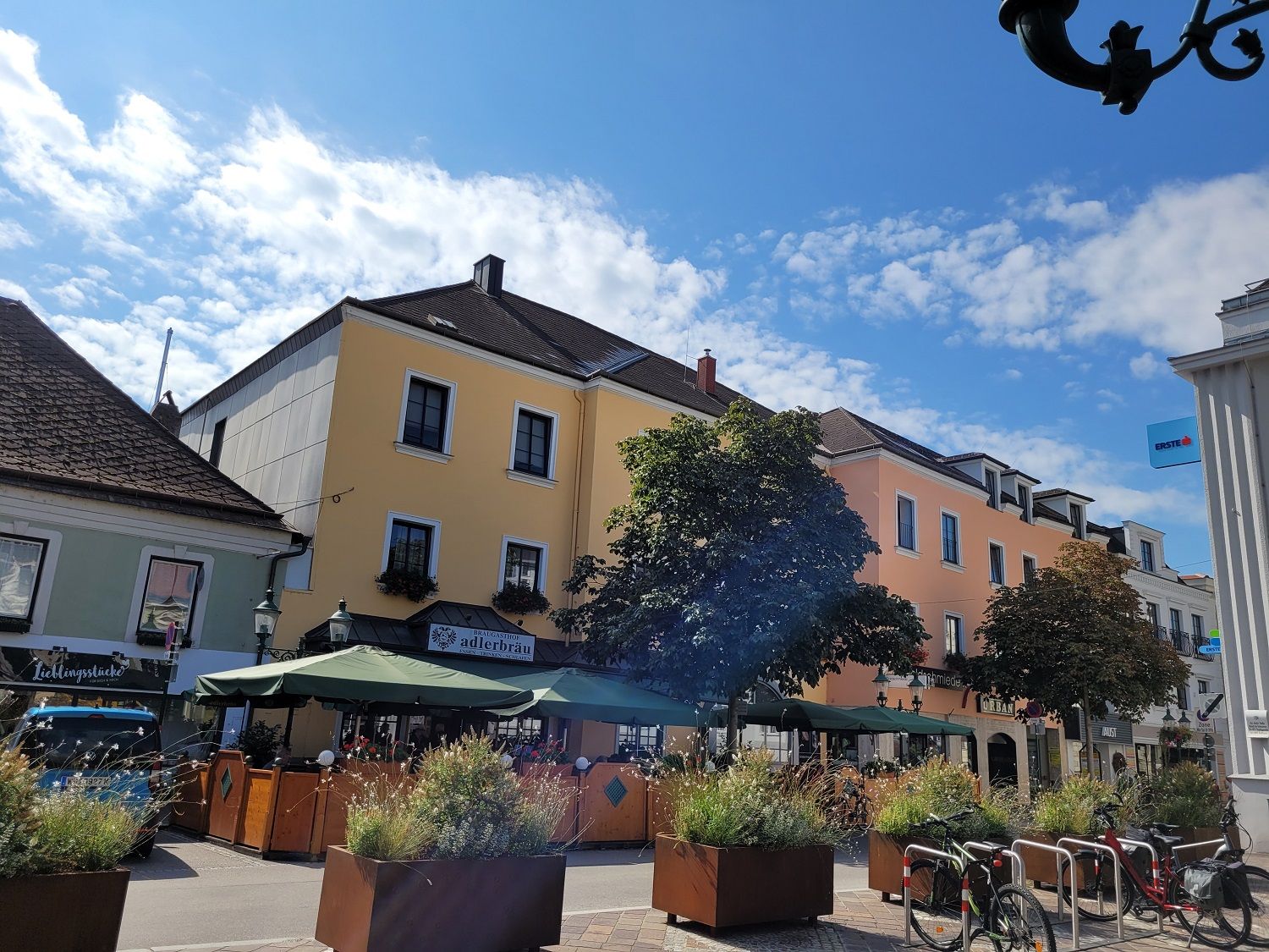 Straßenszene mit gelbem Gebäude, Café mit Sonnenschirmen und Fahrrädern im Vordergrund.