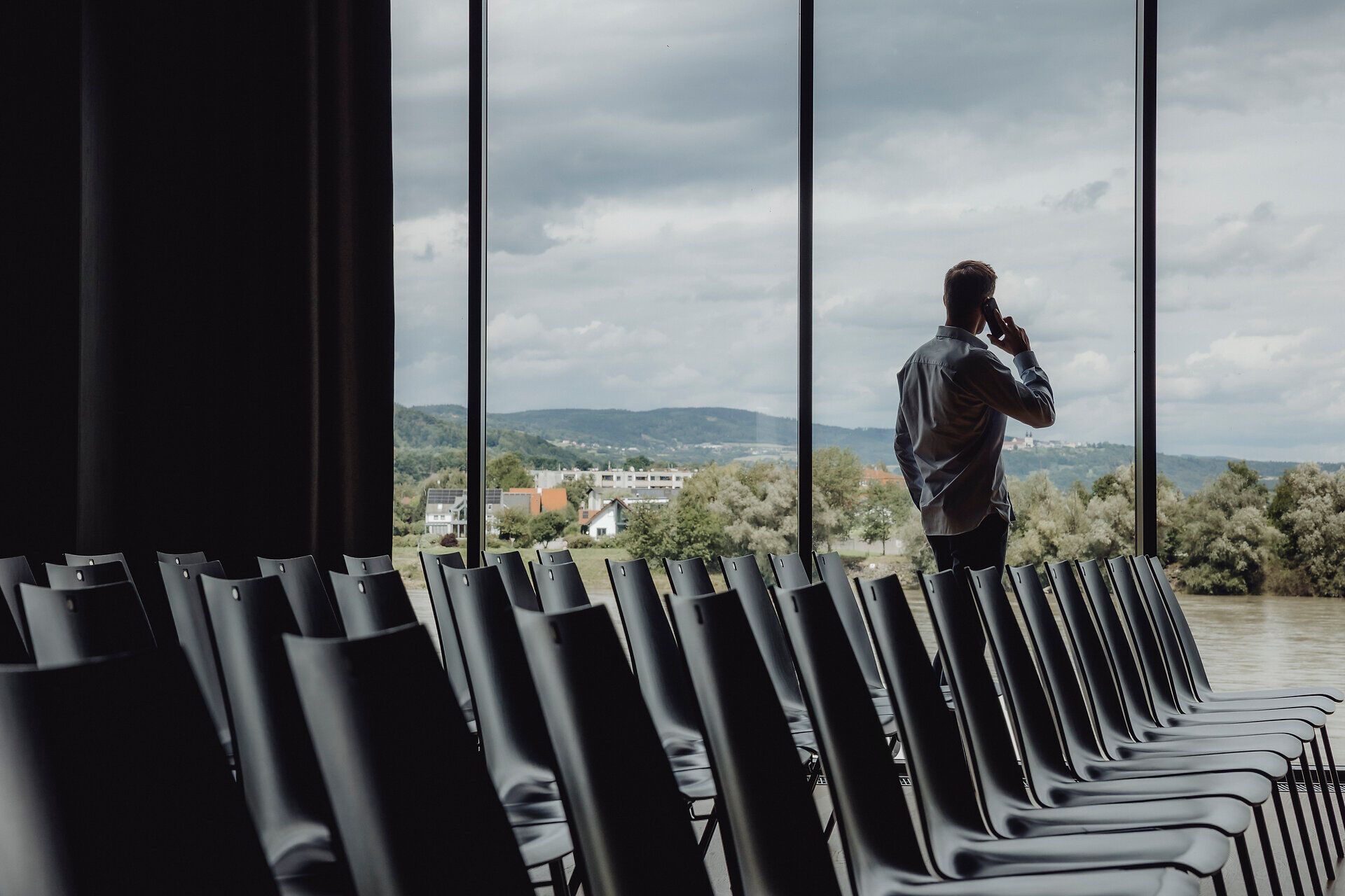 In der Stadthalle Ybbs, umgeben von großen Fenstern, die den Blick auf die sanften Hügel der Donau eröffnen, herrscht eine ruhige, nachdenkliche Atmosphäre. Ein Besucher telefoniert, während die leeren Stühle auf die kommenden Veranstaltungen warten und die Wolken am Himmel eine dramatische Kulisse bieten.