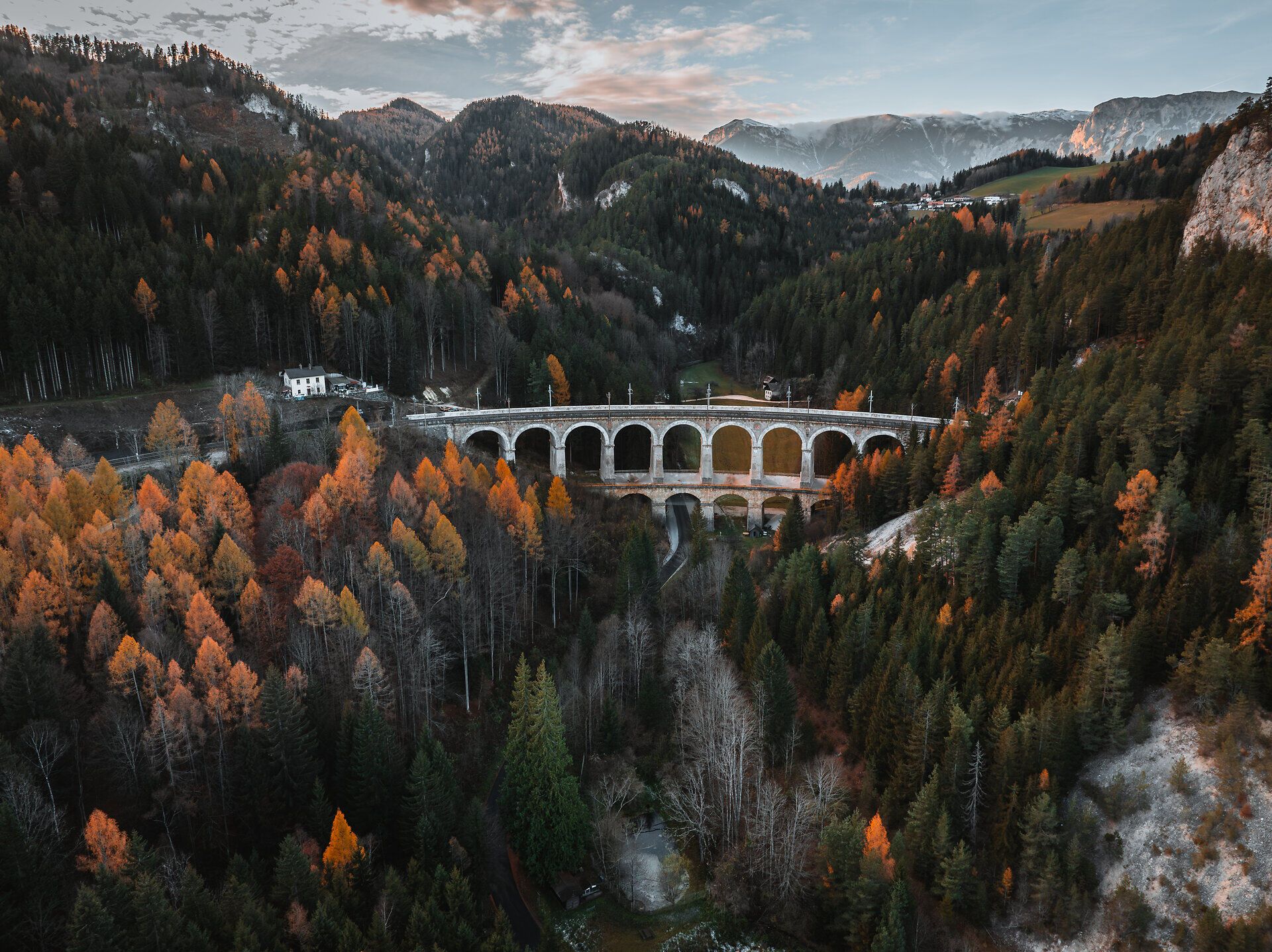Die majestätische Semmeringbahn schlängelt sich durch die bunten Herbstwälder der Wiener Alpen. Goldene und rote Blätter umrahmen die beeindruckenden Bögen der Brücke, während die sanften Hügel im Hintergrund eine malerische Kulisse bieten. Ein Ort, der die Seele berührt und zum Verweilen einlädt.