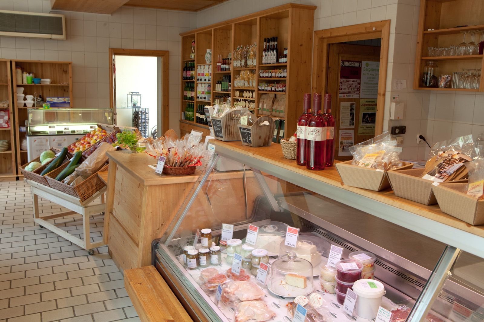 Innenansicht eines Feinkostladens mit Holzregalen, Gemüse, Weinflaschen und einer Kühltheke mit Käse und Aufstrichen.