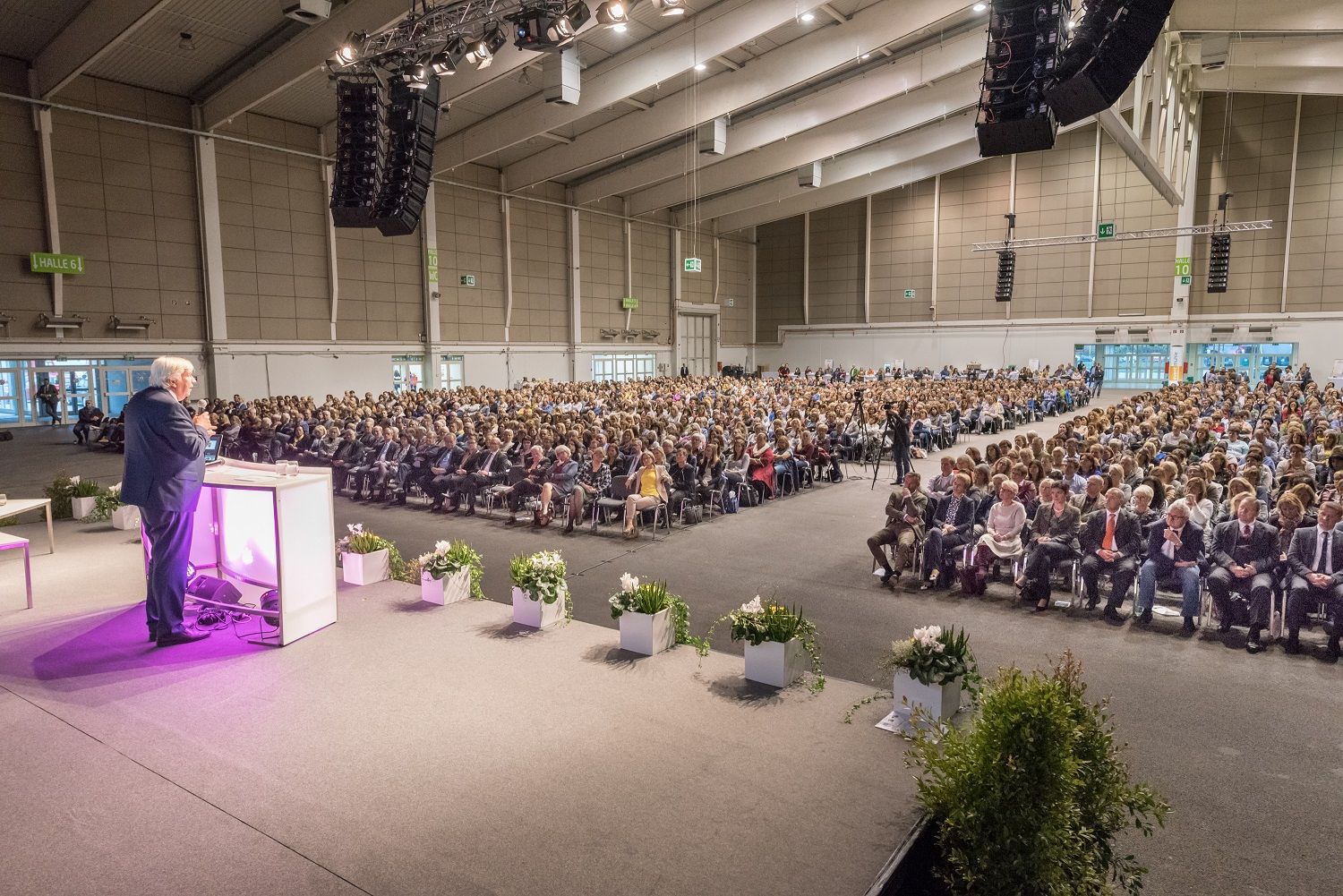 Ein Redner spricht vor einem großen Publikum in einer Messehalle.