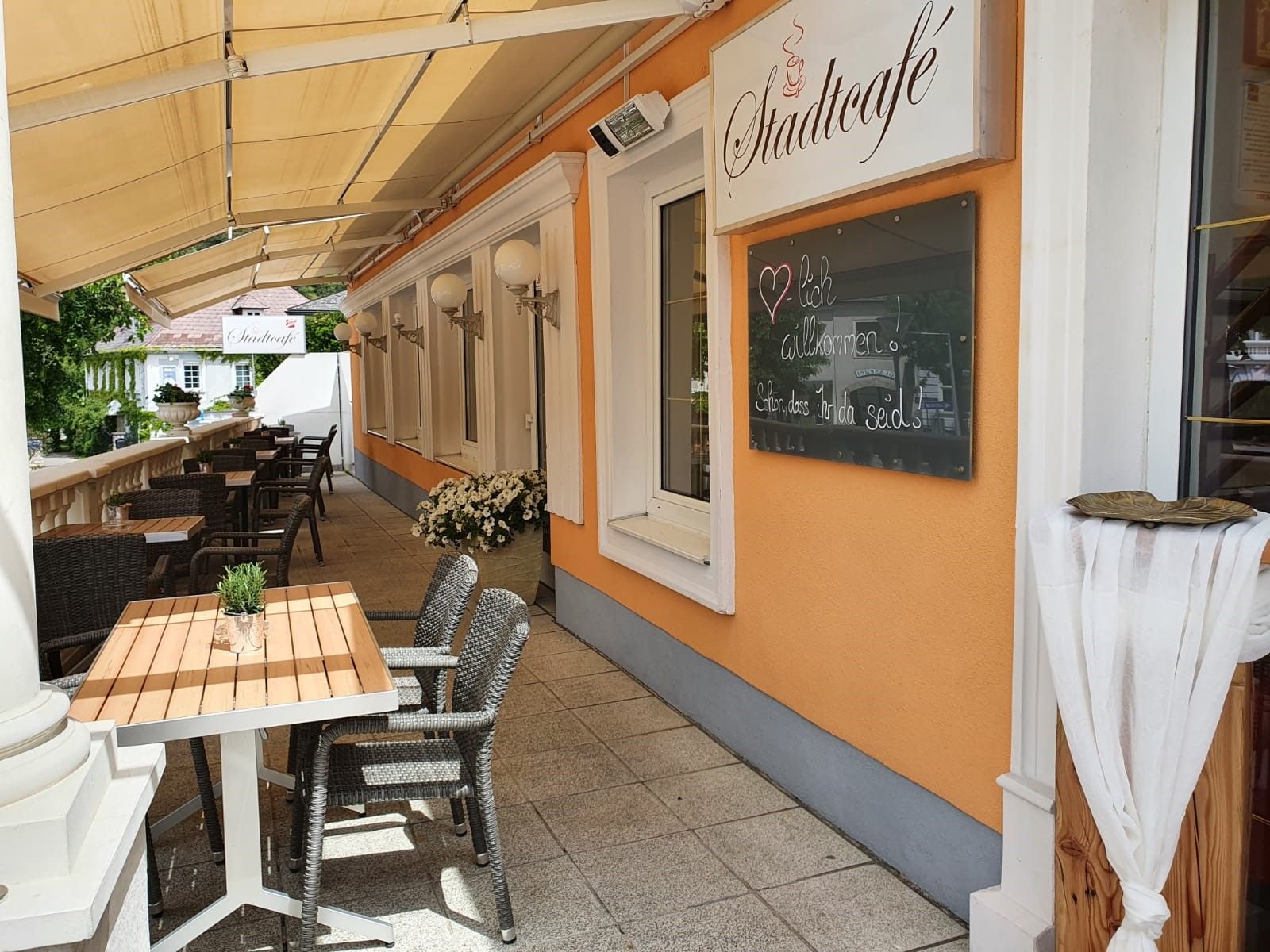 Außenbereich eines Cafés mit Tischen und Stühlen, orangefarbene Wand mit Schild 'Stadtcafé'.