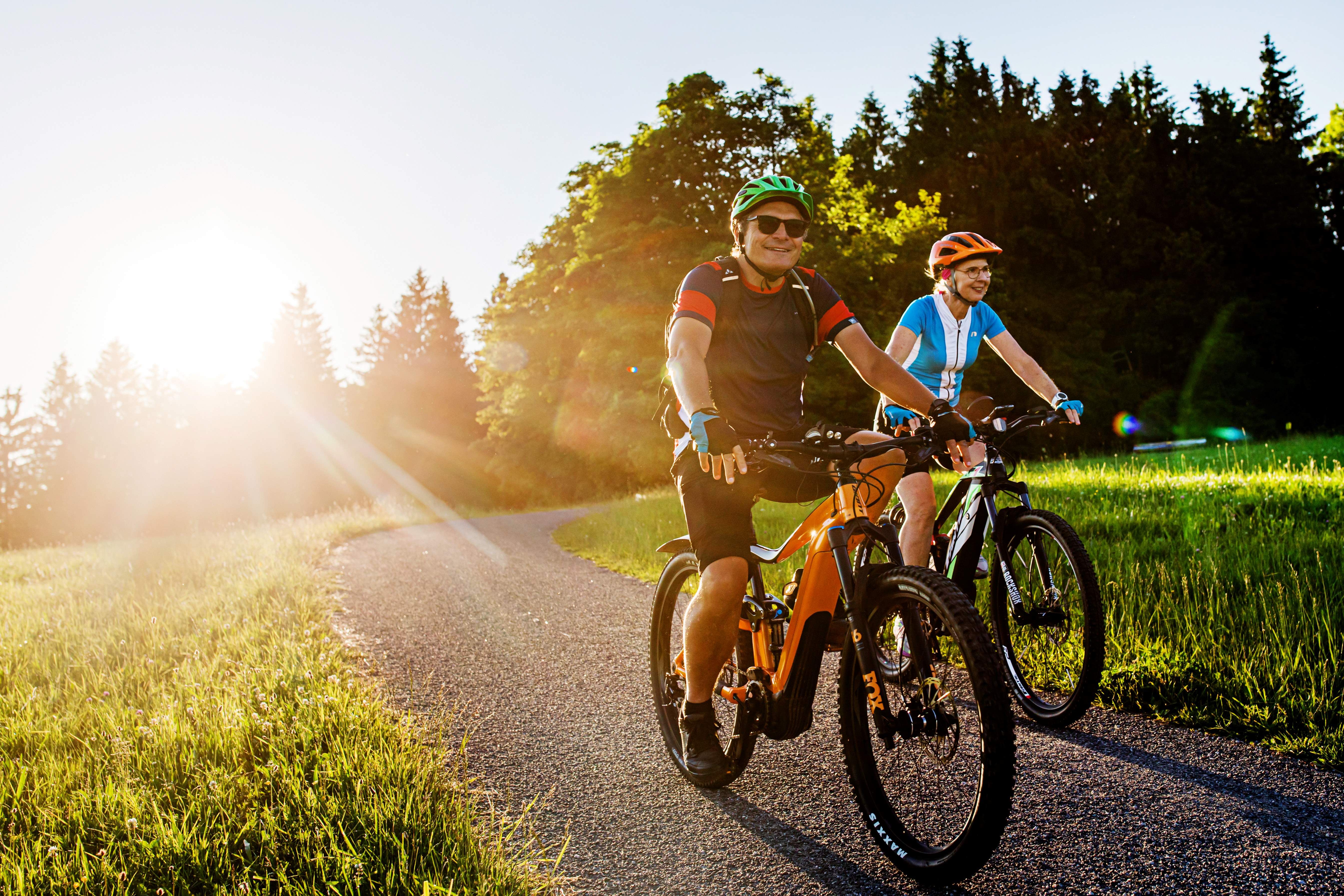 Zwei Personen fahren bei Sonnenuntergang auf E-Bikes auf einem Weg durch eine grüne Landschaft.