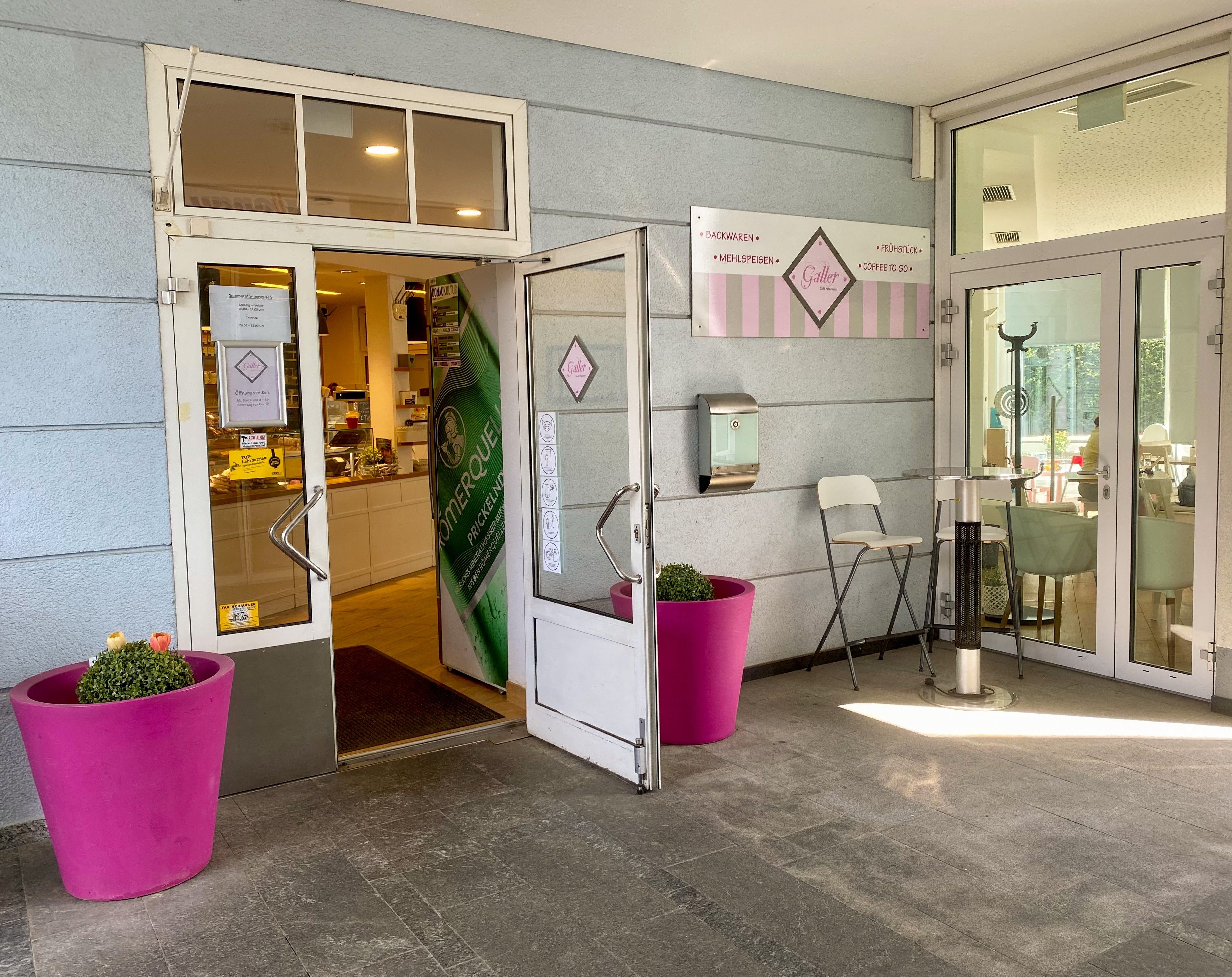 Eingang Kaffeehaus klinks und rechts rosa Blumentöpfe vor hellblauem Haus 