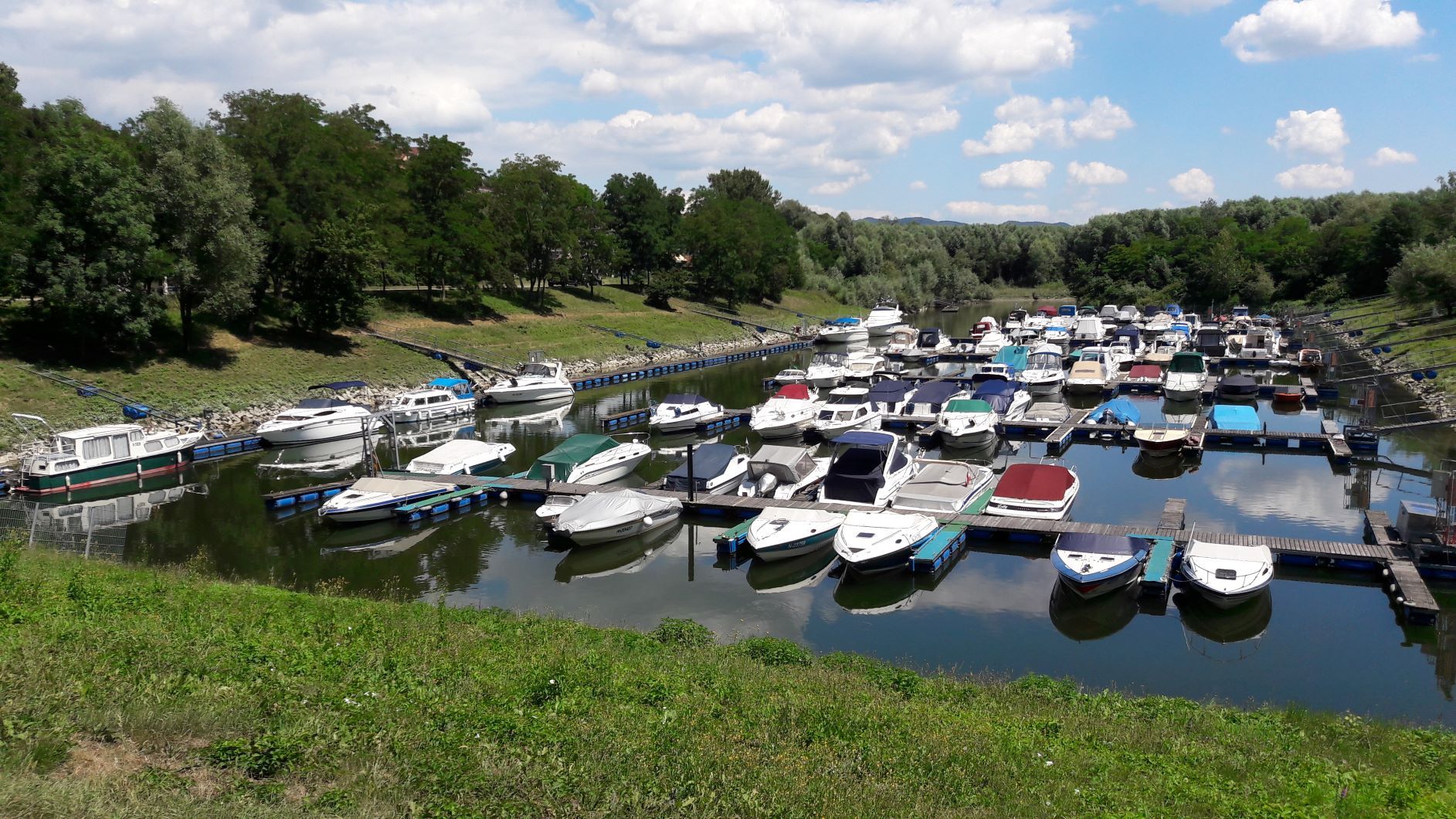 Boote im Sportboothafen Emmersdorf, umgeben von grünen Bäumen und blauem Himmel.