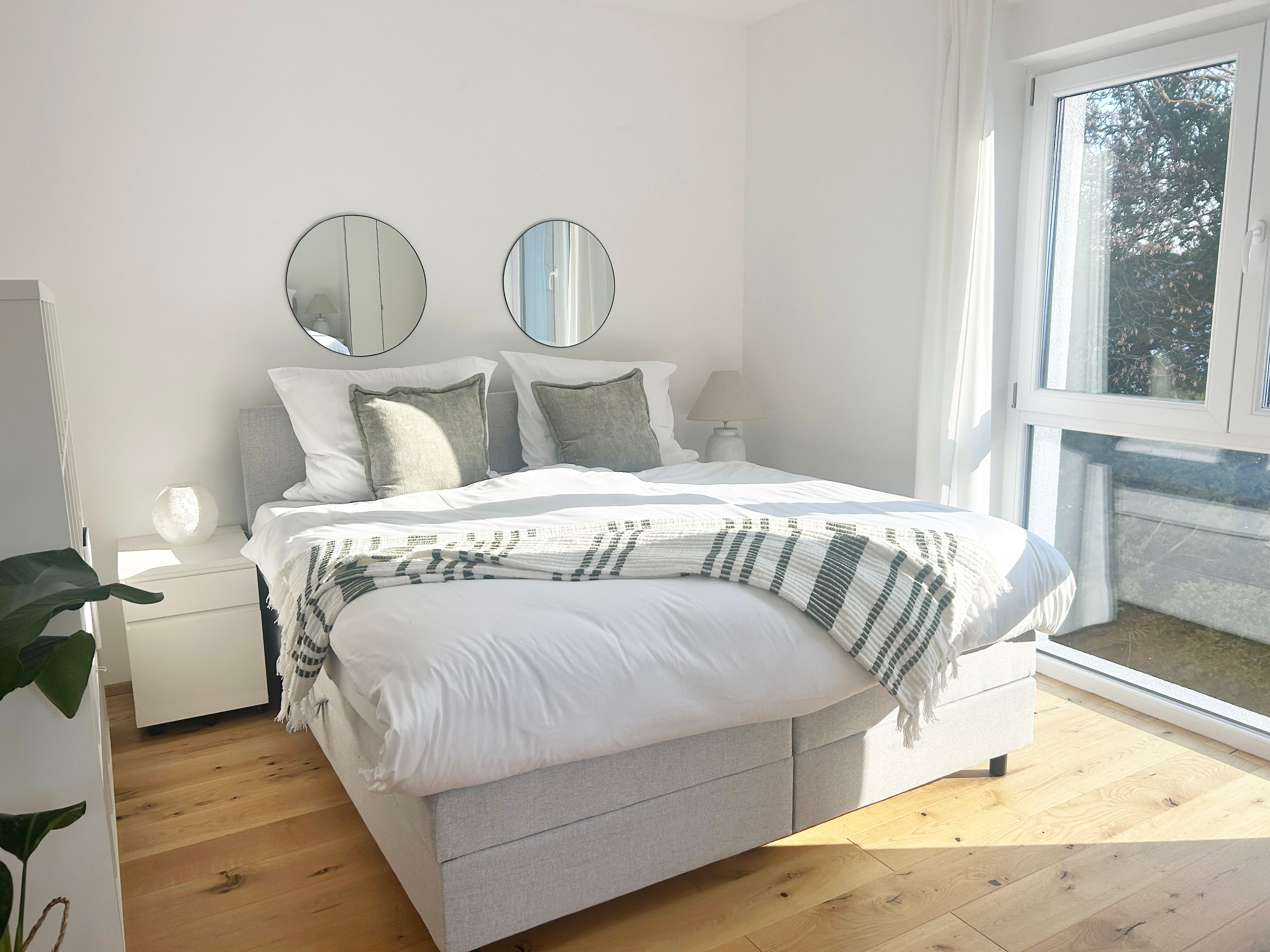Helles Schlafzimmer mit Doppelbett, zwei runden Spiegeln an der Wand und großen Fenstern.