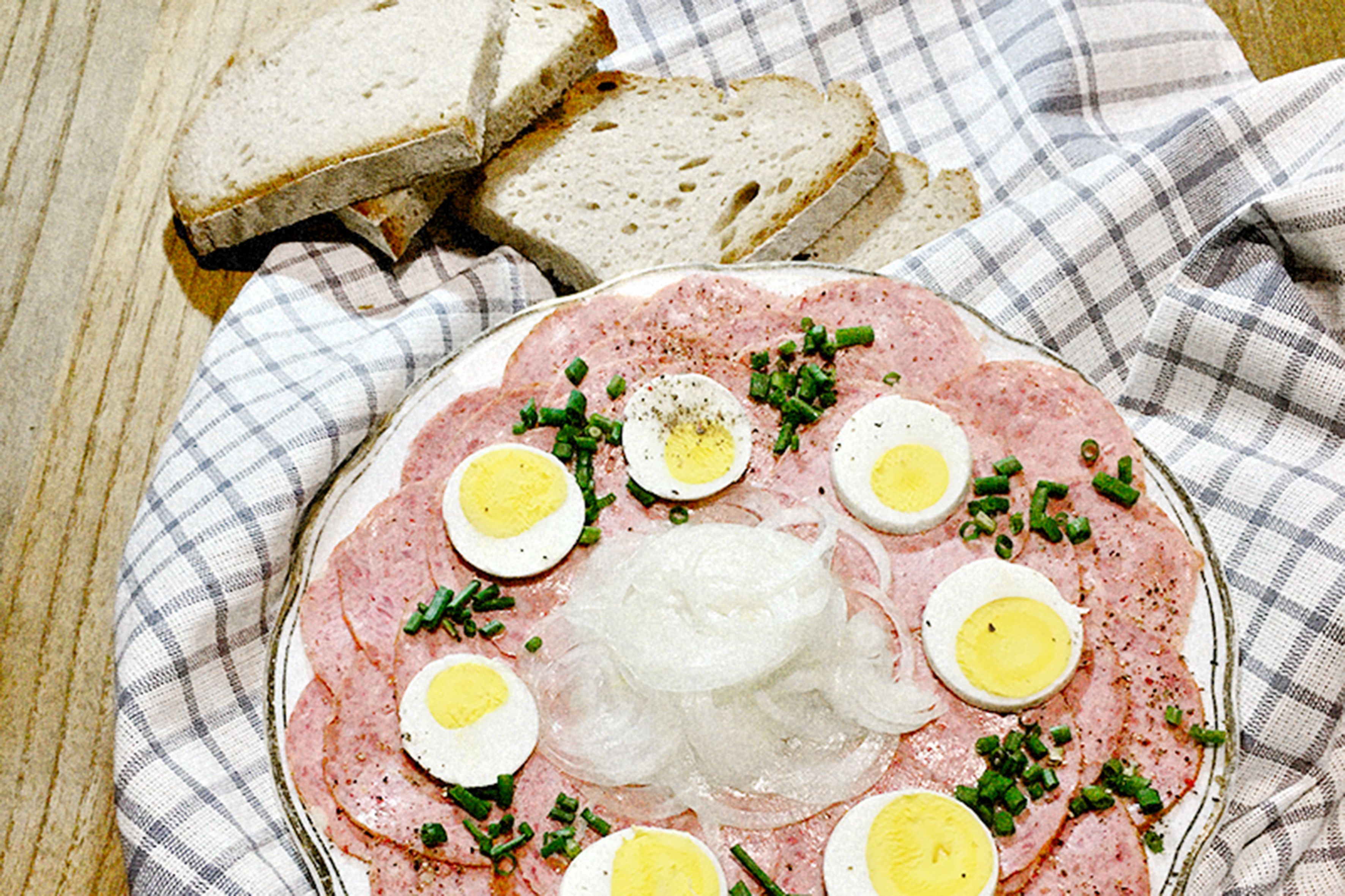 Ein Teller mit Wurstscheiben, gekochten Eiern und Zwiebeln, garniert mit Schnittlauch, daneben Brotscheiben auf einem karierten Tuch.