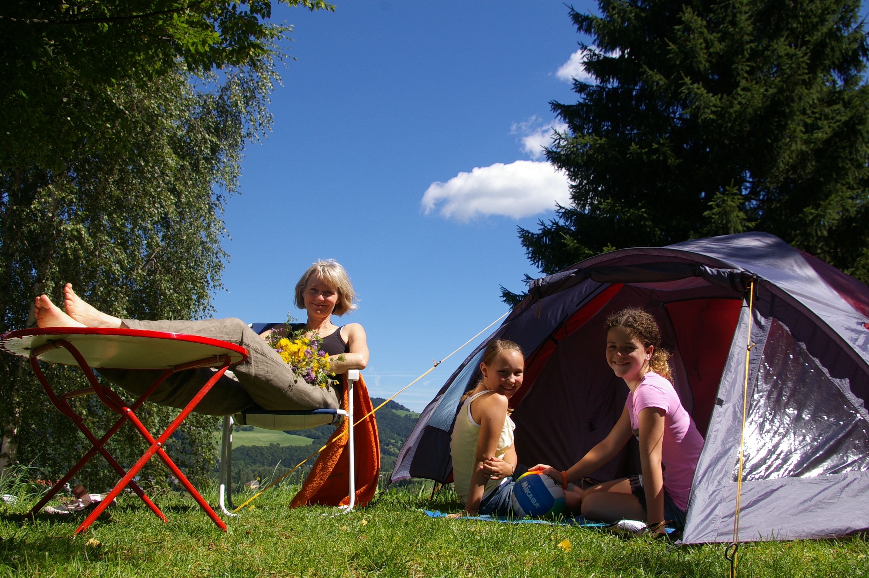 Eine Frau entspannt sich auf einem Campingstuhl neben einem Zelt, während zwei Kinder im Zelt sitzen. Im Hintergrund sind Bäume und blauer Himmel zu sehen.