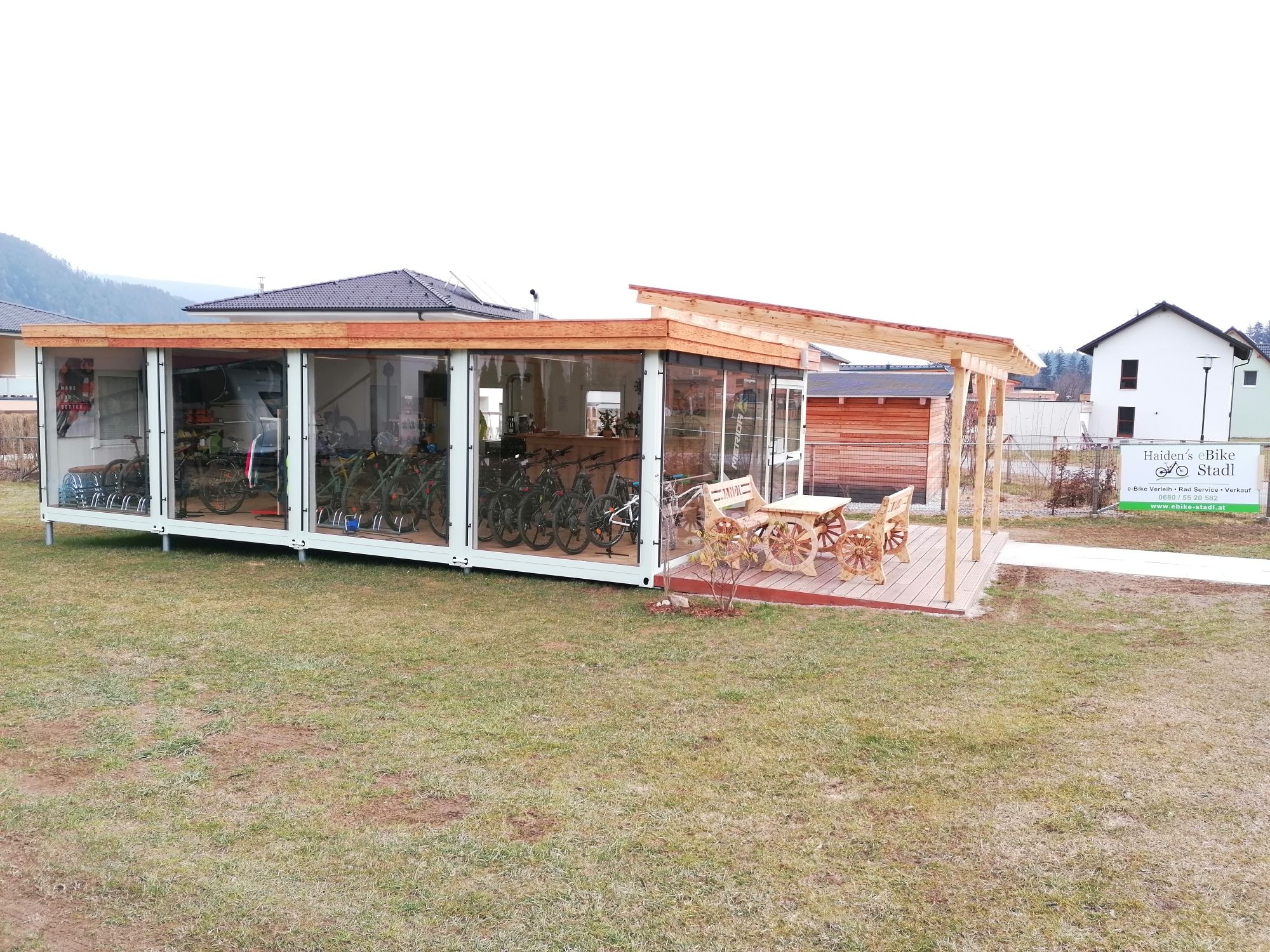 Außenansicht eines modernen Fahrradverleihs mit Glasfront und Holzdach, umgeben von Rasen und einem Schild mit der Aufschrift 'Haiden's eBike Stadl'.