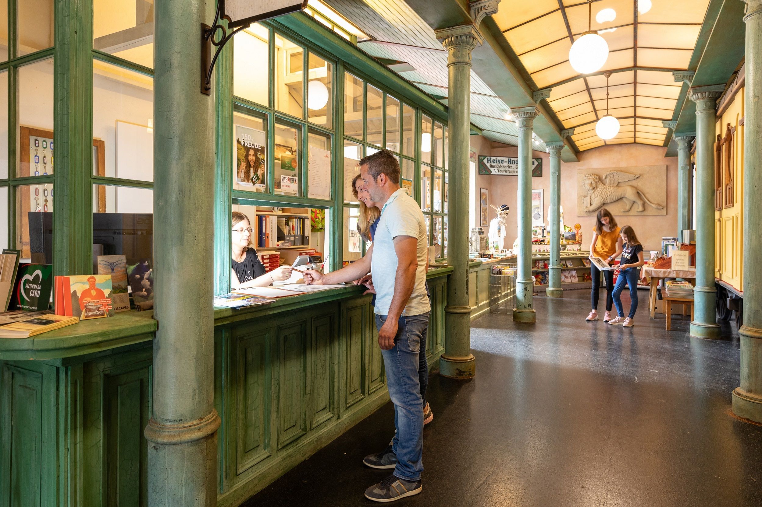 Innenansicht des Südbahn Museums mit Besuchern an einem Schalter und zwei Personen, die ein Buch betrachten.