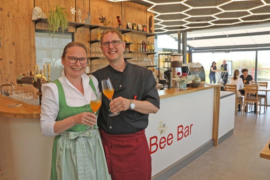 Zwei Personen in traditioneller Kleidung stehen lächelnd vor der Bee Bar mit Getränken in der Hand.
