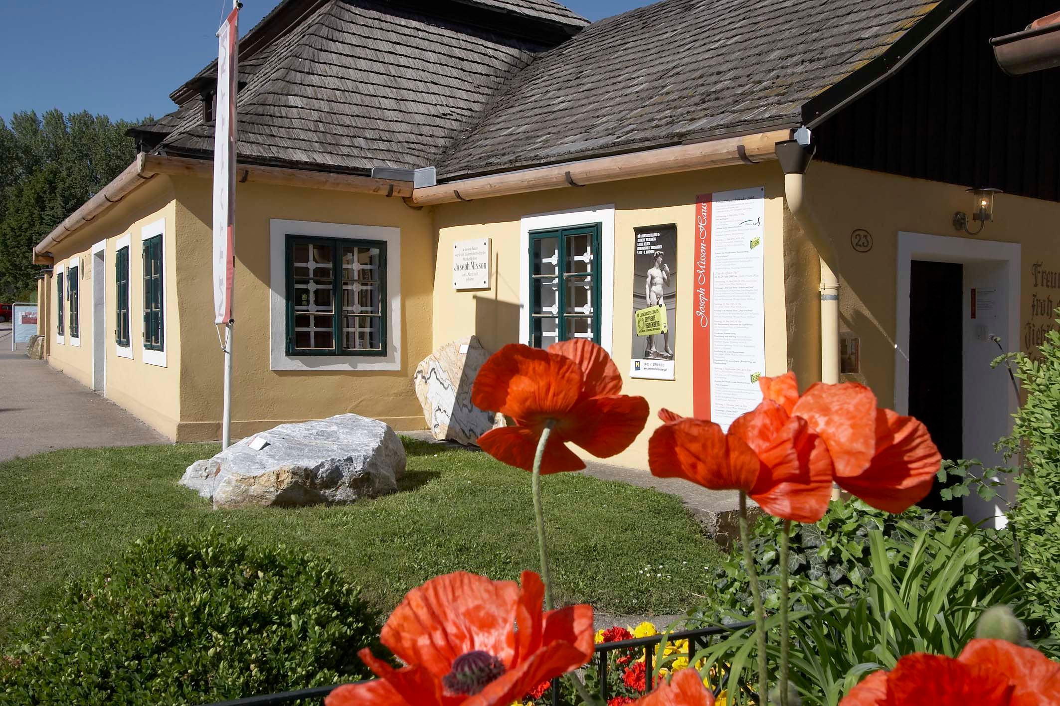 Gelbes Haus mit Schindeldach und Mohnblumen im Vordergrund.
