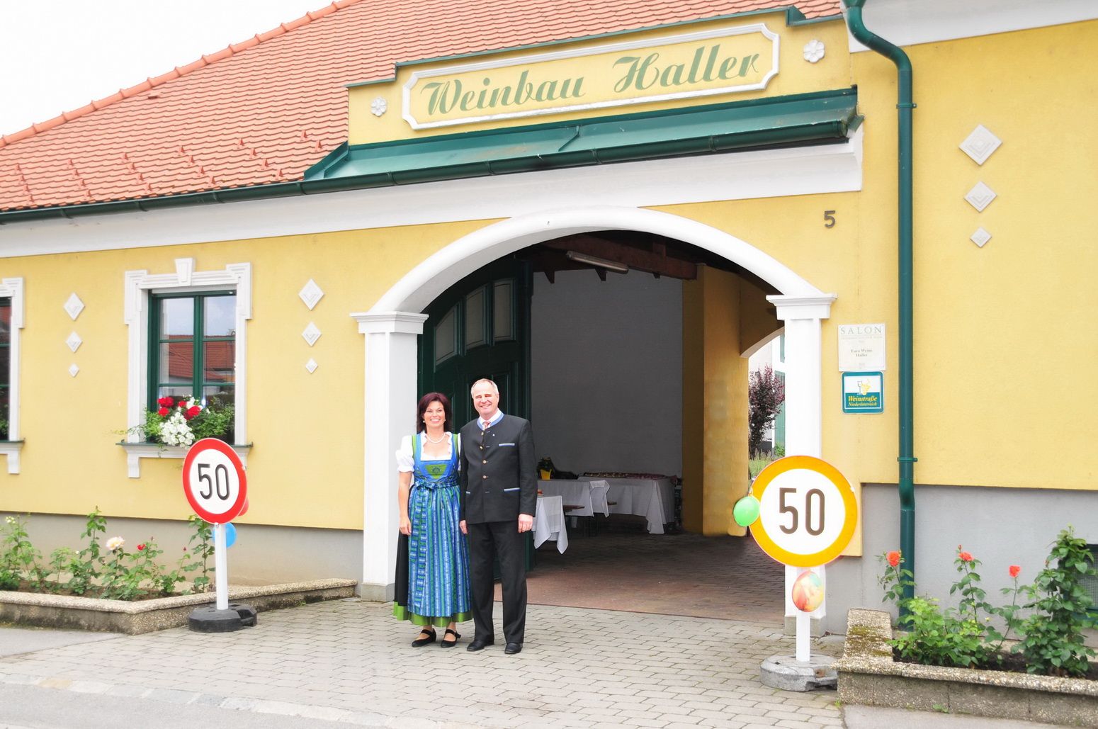 Ein Paar steht vor einem gelben Gebäude mit der Aufschrift 'Weinbau Haller'.
