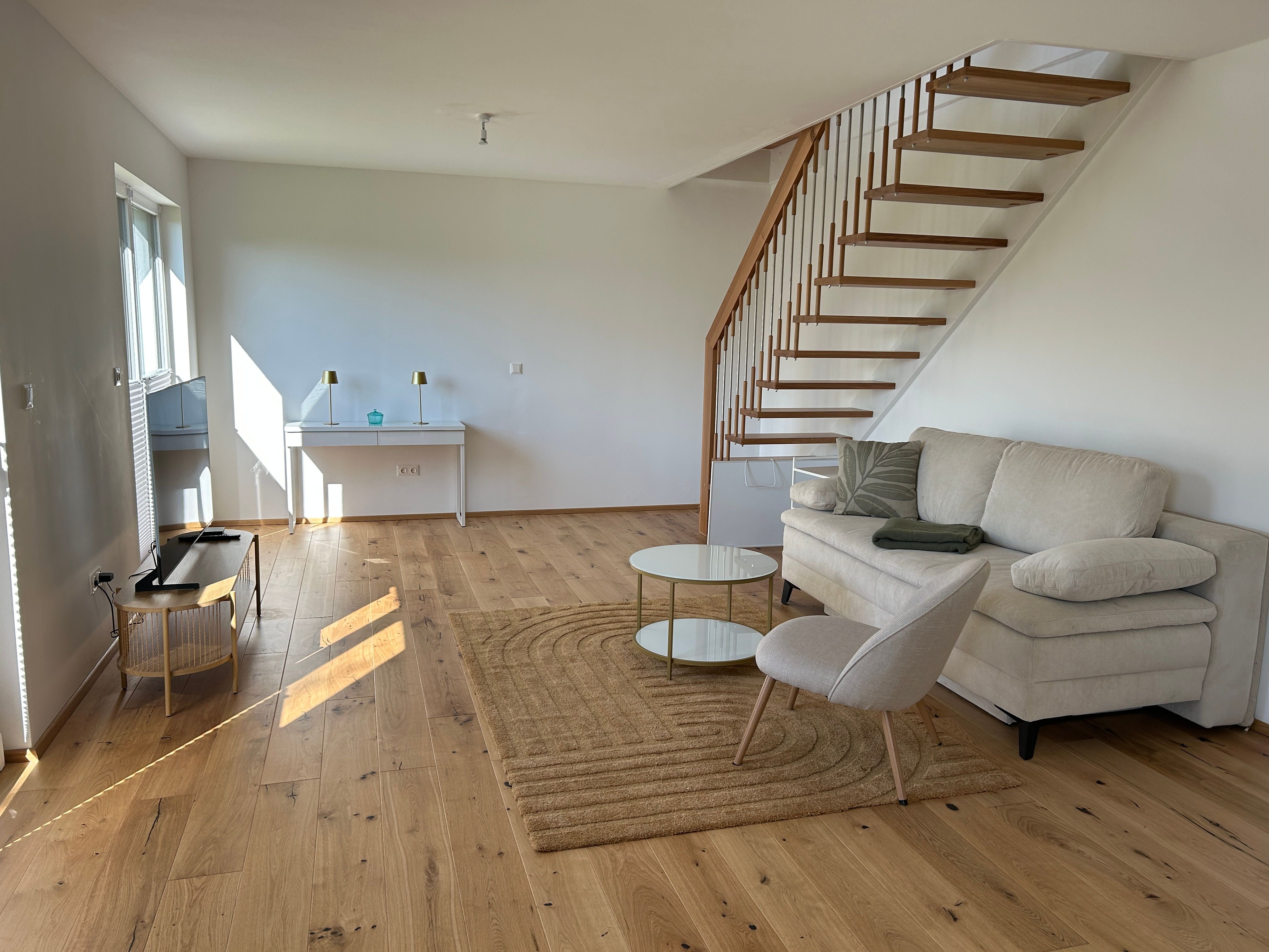 Modernes Wohnzimmer mit Holztreppe, Sofa, Sessel und Couchtisch auf Holzboden.