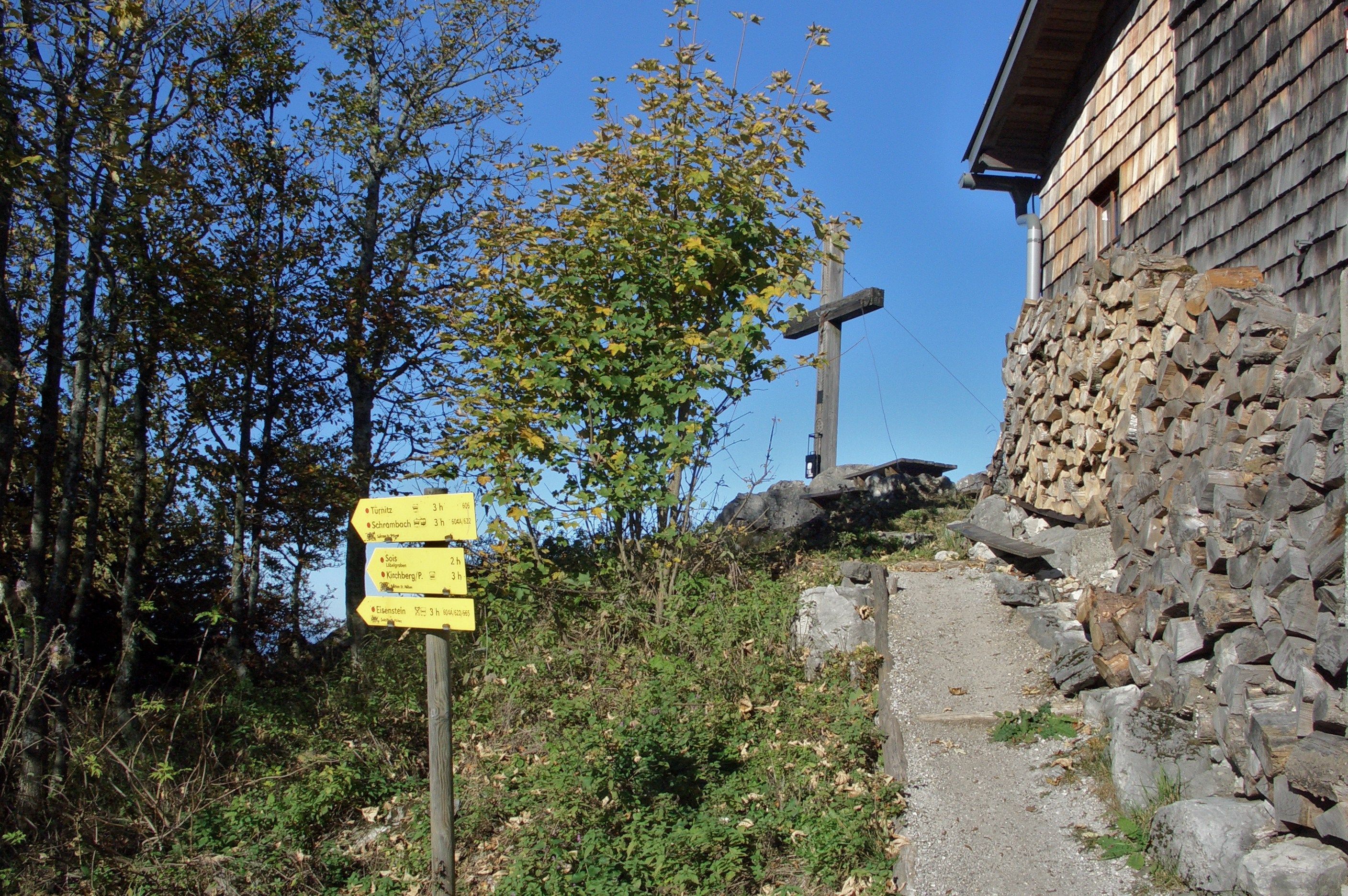 Wegweiser und Holzkreuz neben einem Holzhaus in einer bergigen Landschaft.