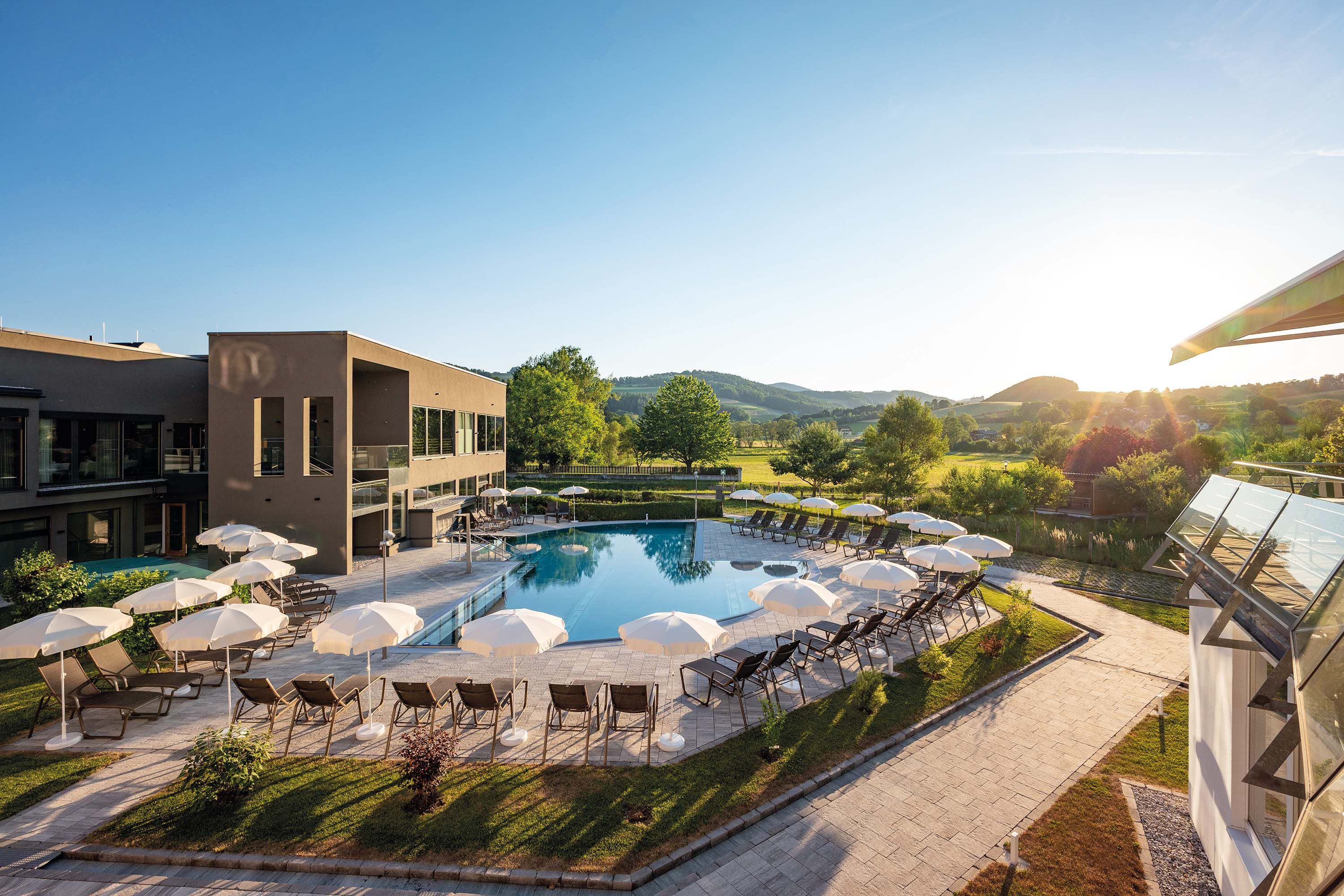 Außenanlage eines Hotels mit Pool, Sonnenschirmen, Liegestühlen, umgeben von grünen Wiesen und Bäumen.