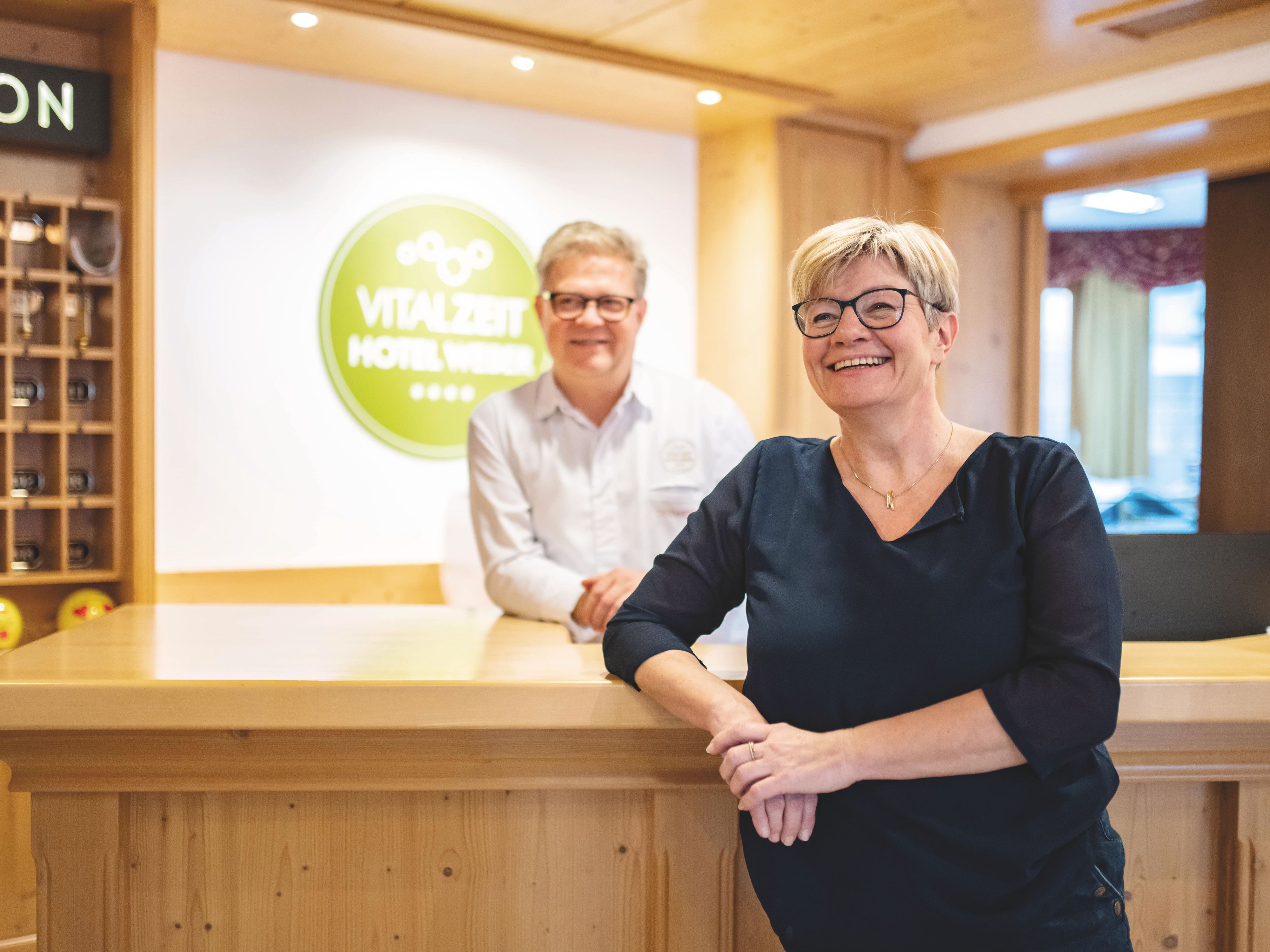 Petra und Alfred Weber stehen lächelnd an der Rezeption des VitalZeit Hotel Weber.