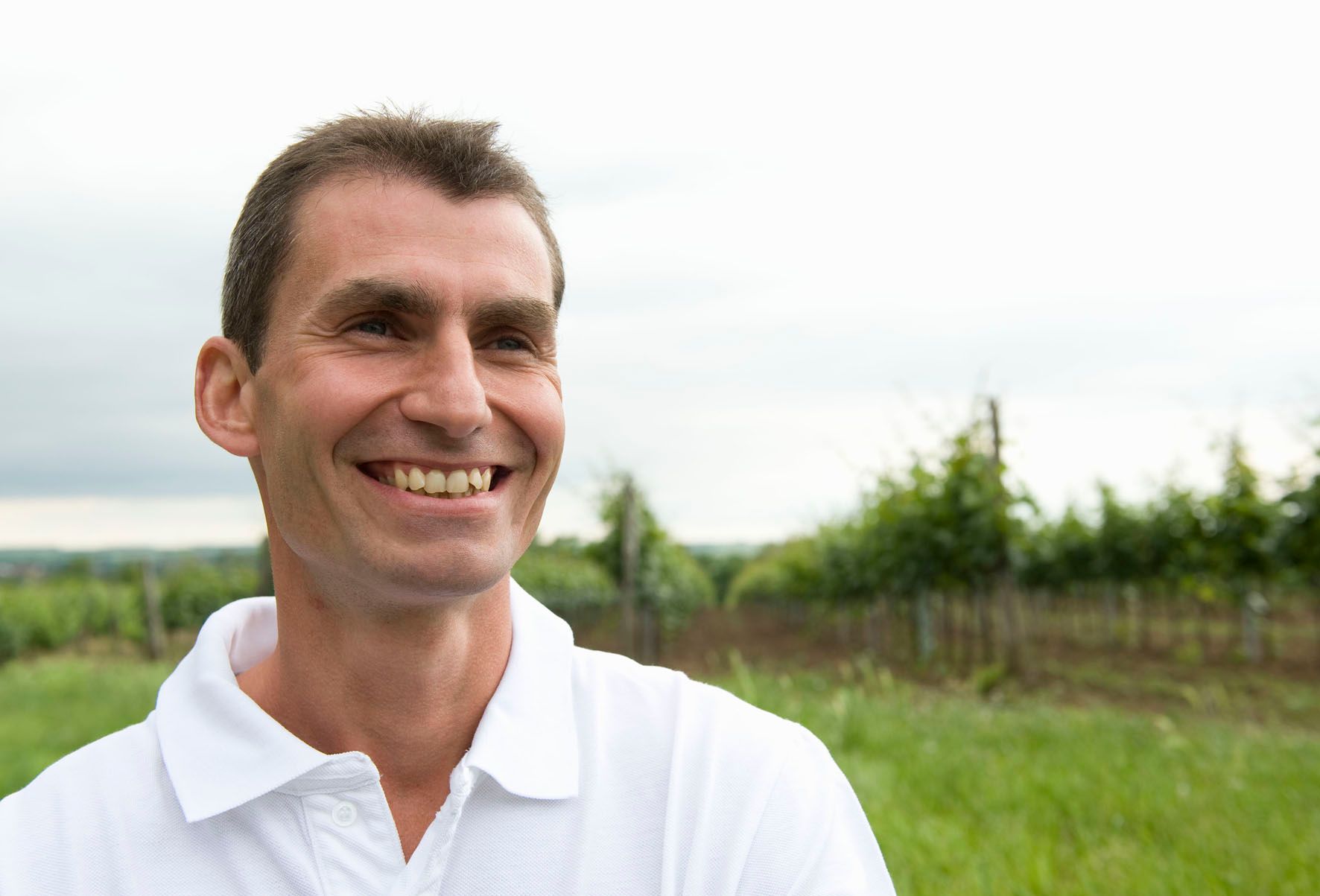 Ein lächelnder Mann in einem weißen Polohemd steht auf einem Feld mit Bäumen im Hintergrund.