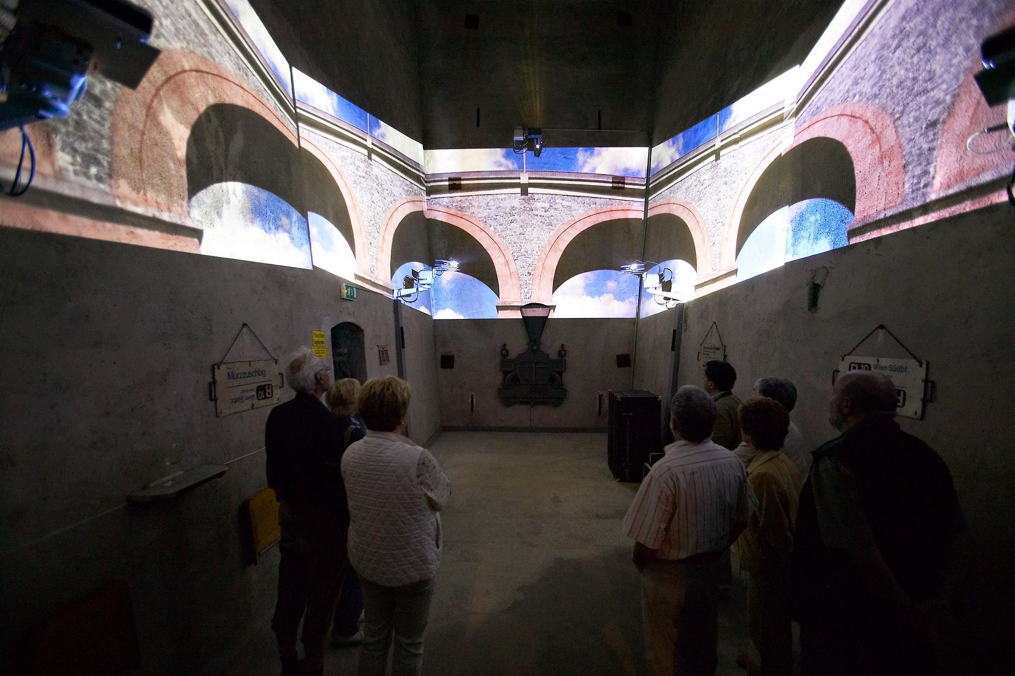 Menschen betrachten eine Multimedia-Show in einem Raum mit Projektionen von Bögen an den Wänden.