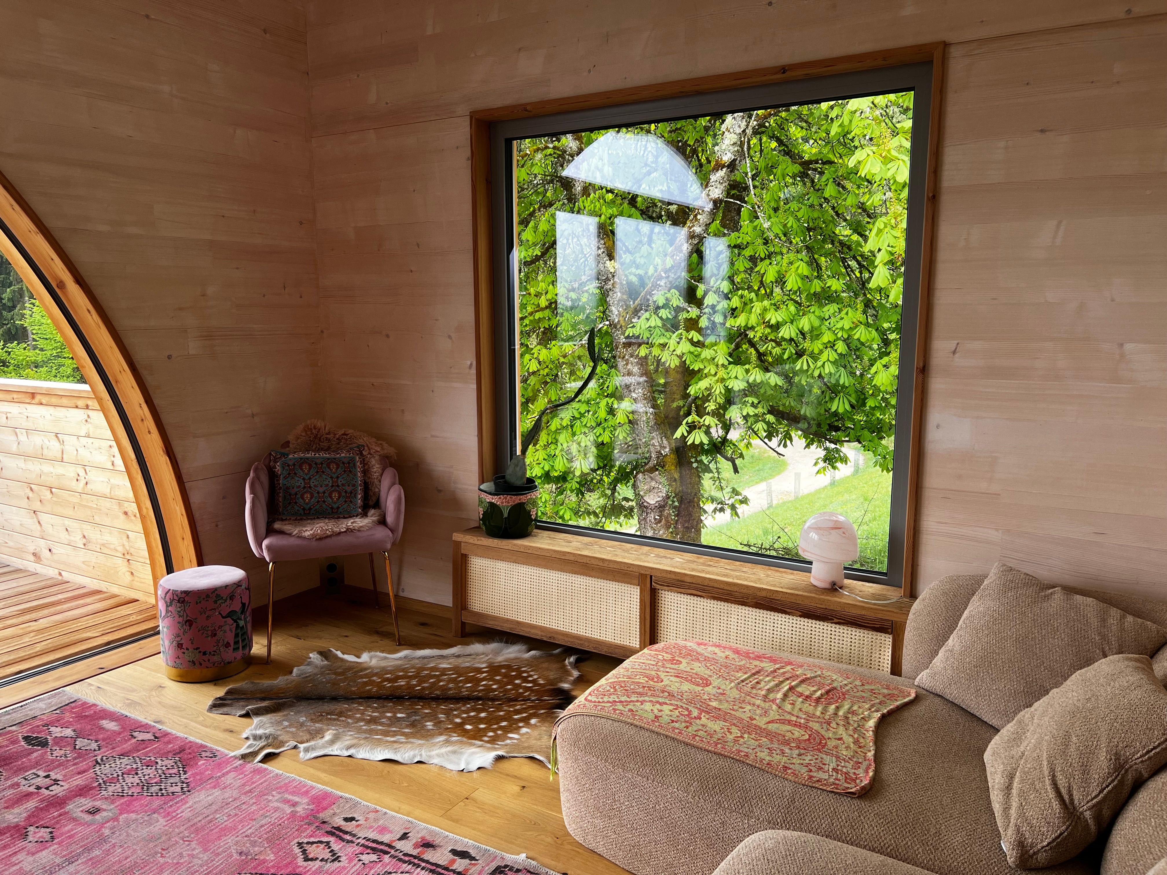 Innenansicht eines gemütlichen Raumes mit großem Fenster, das auf einen grünen Baum blickt. Ein Sessel, ein Hocker und ein Sofa sind zu sehen.