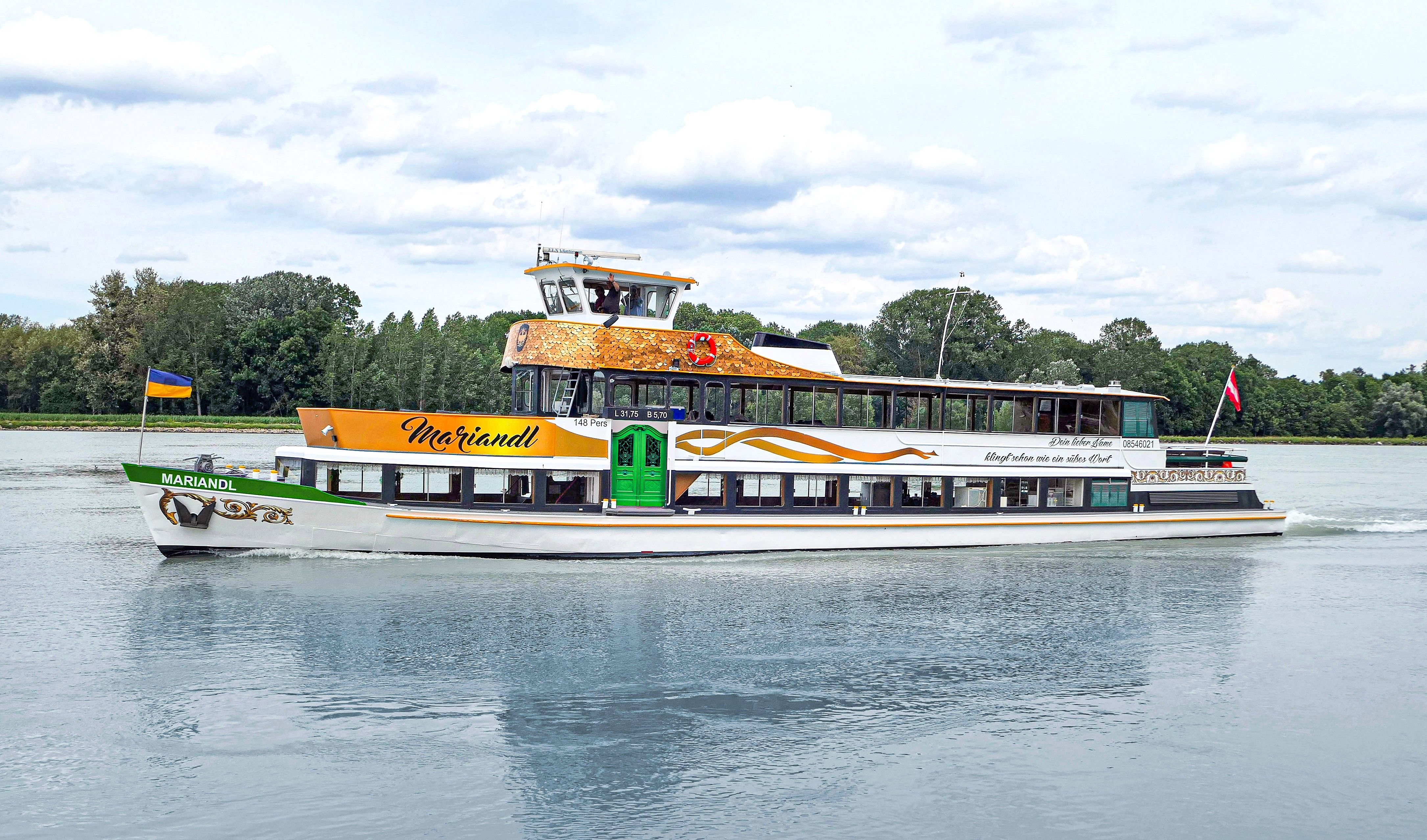 Ein Ausflugsschiff namens MS Mariandl fährt auf einem Fluss, umgeben von Bäumen. Es trägt eine ukrainische und eine österreichische Flagge.