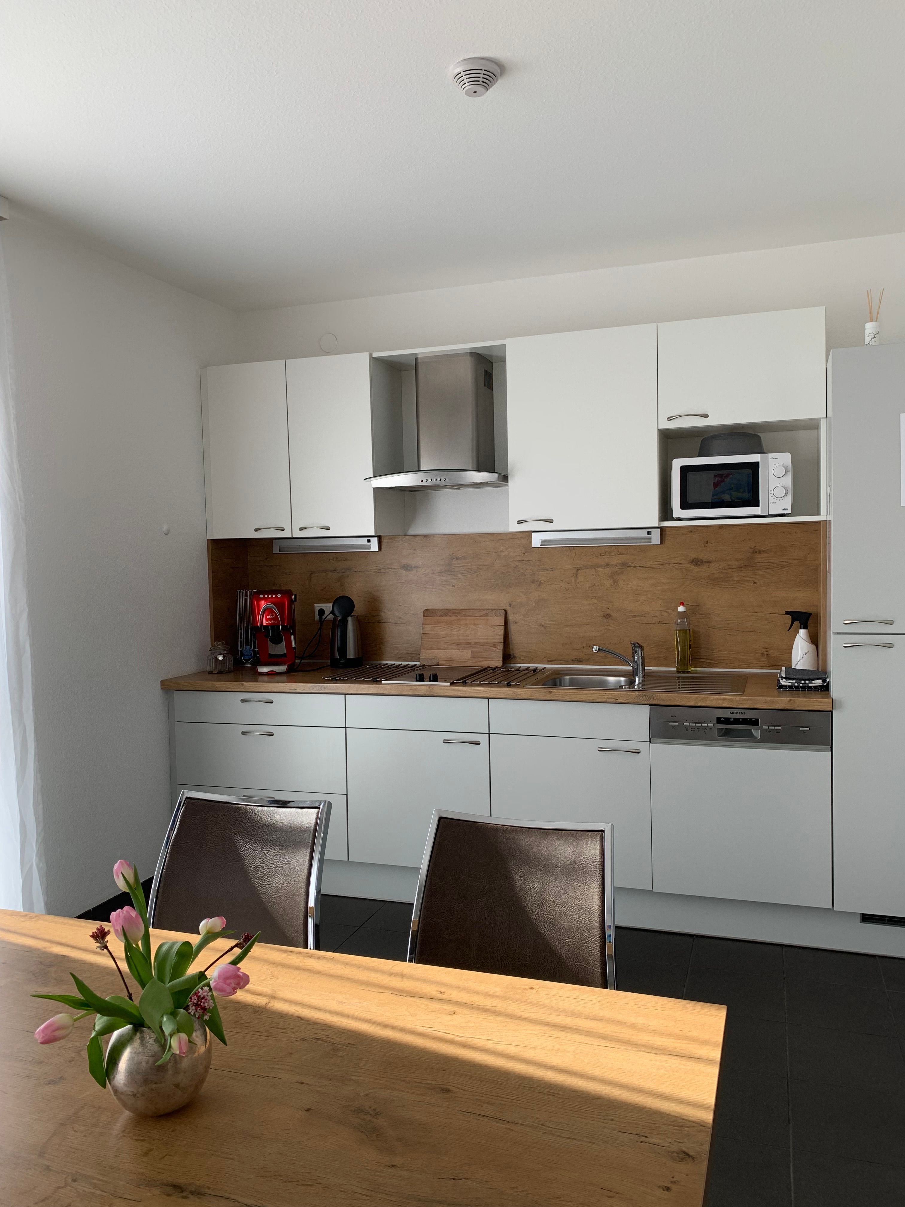 Moderne Küchenzeile mit weißen Schränken, Holzarbeitsplatte, Kaffeemaschine und Mikrowelle. Ein Tisch mit Blumenstrauß im Vordergrund.