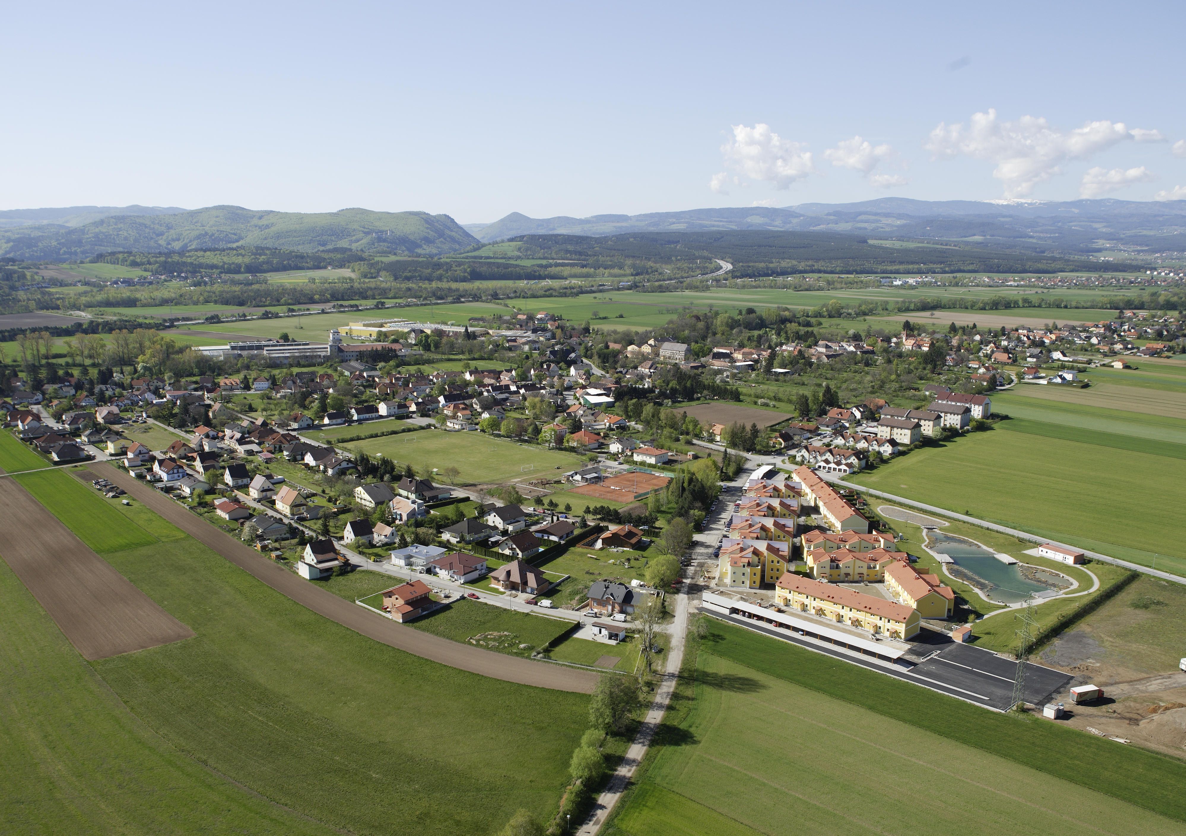 Luftaufnahme eines Dorfes mit Feldern und Hügeln im Hintergrund.