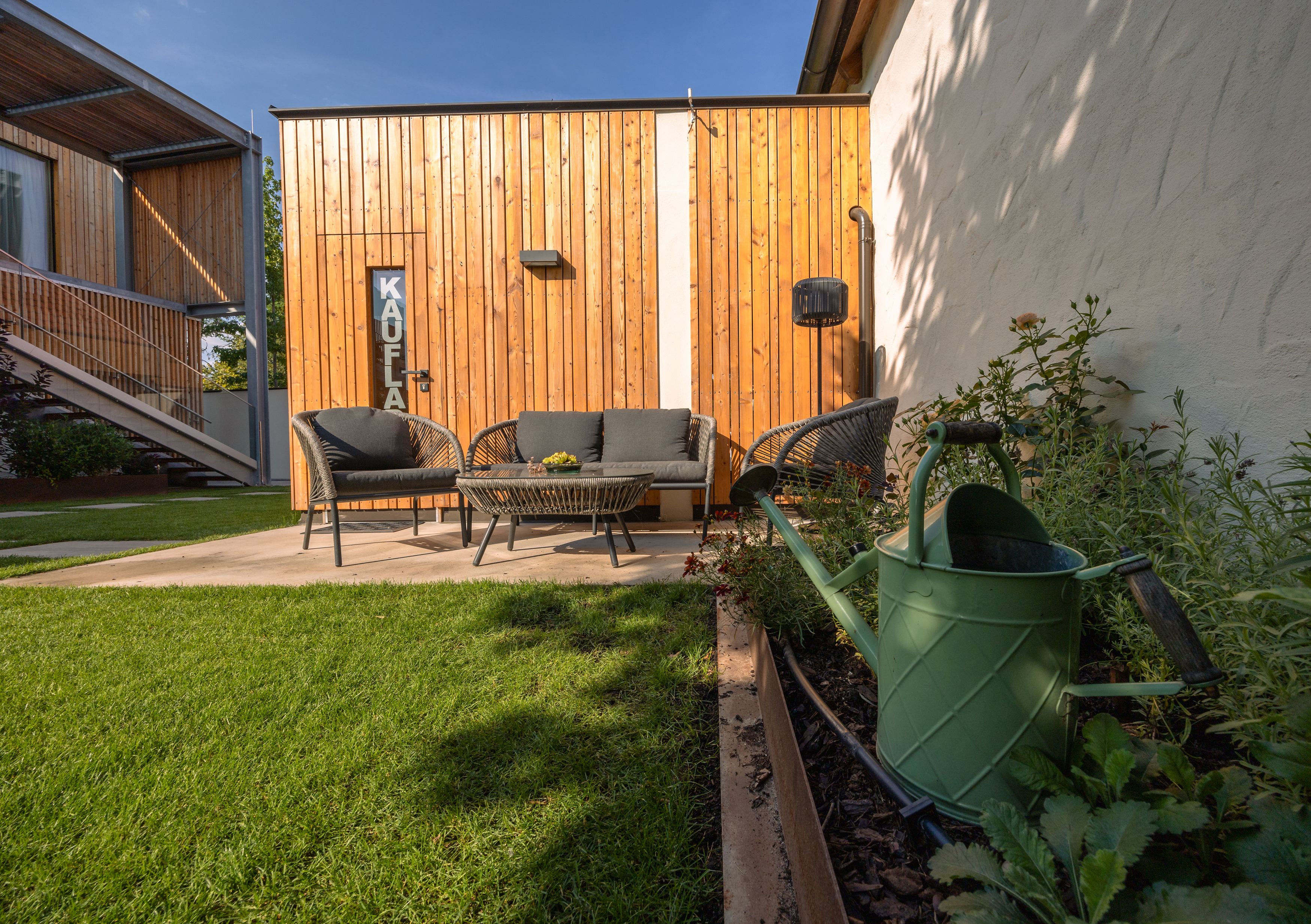 Gartenbereich mit Sitzmöbeln vor einem Holzhaus, Gießkanne im Vordergrund.