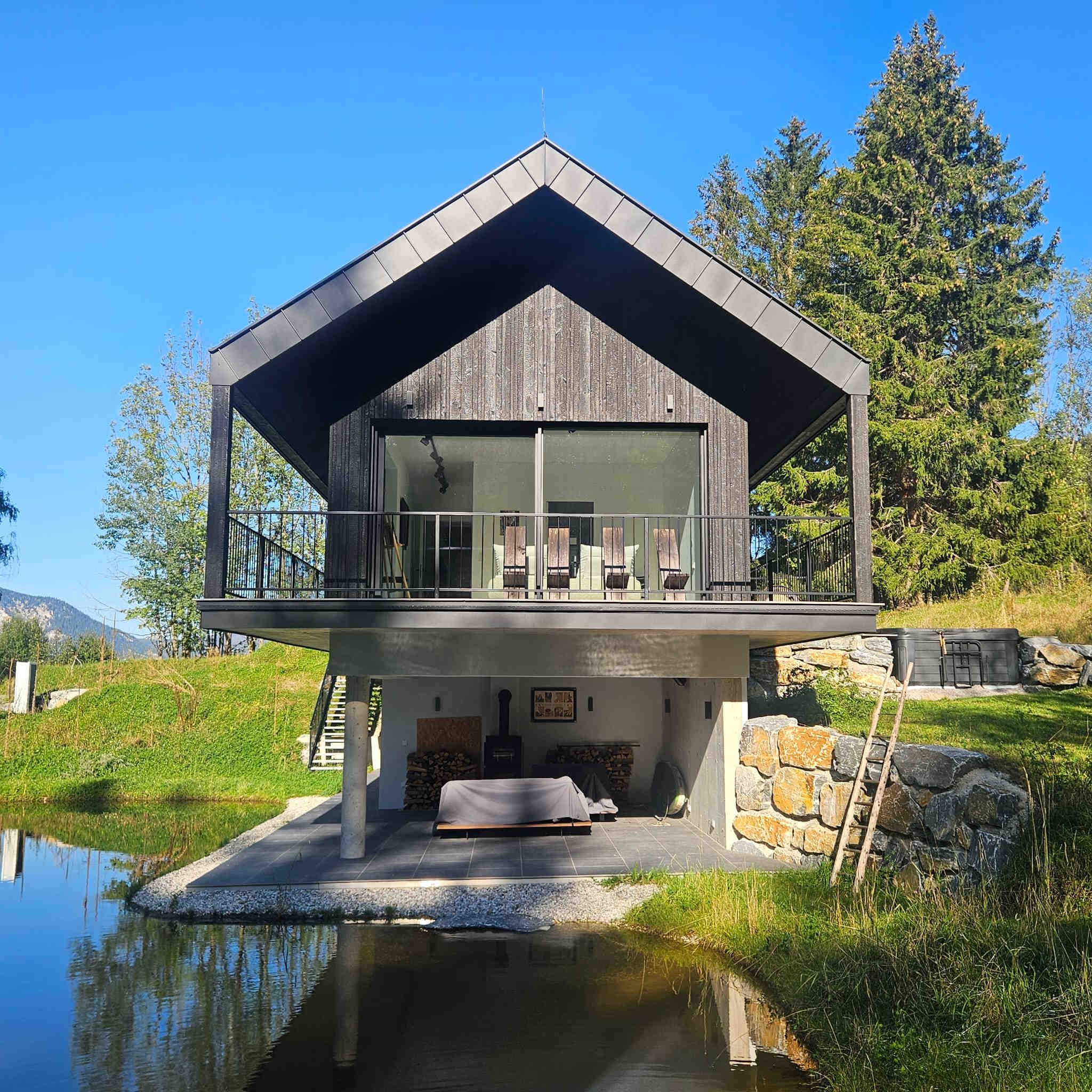 Modernes Haus mit Holzfassade und Balkon, umgeben von Natur und einem kleinen Teich.