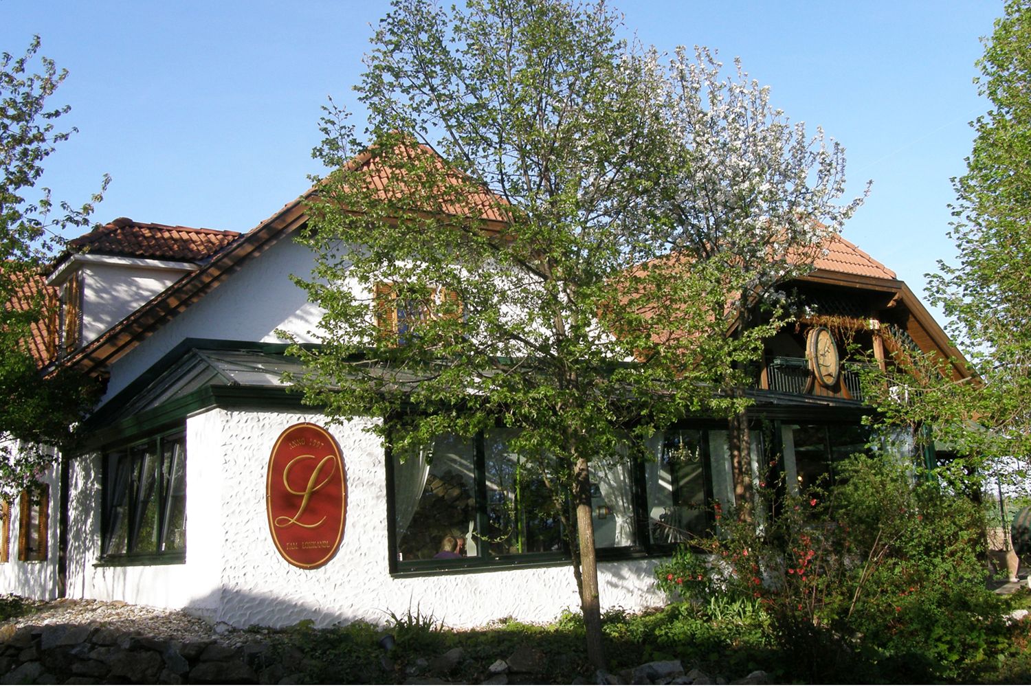 Außenansicht eines traditionellen Gebäudes mit roten Dachziegeln und einem großen L-Logo an der Wand, umgeben von Bäumen und Pflanzen.