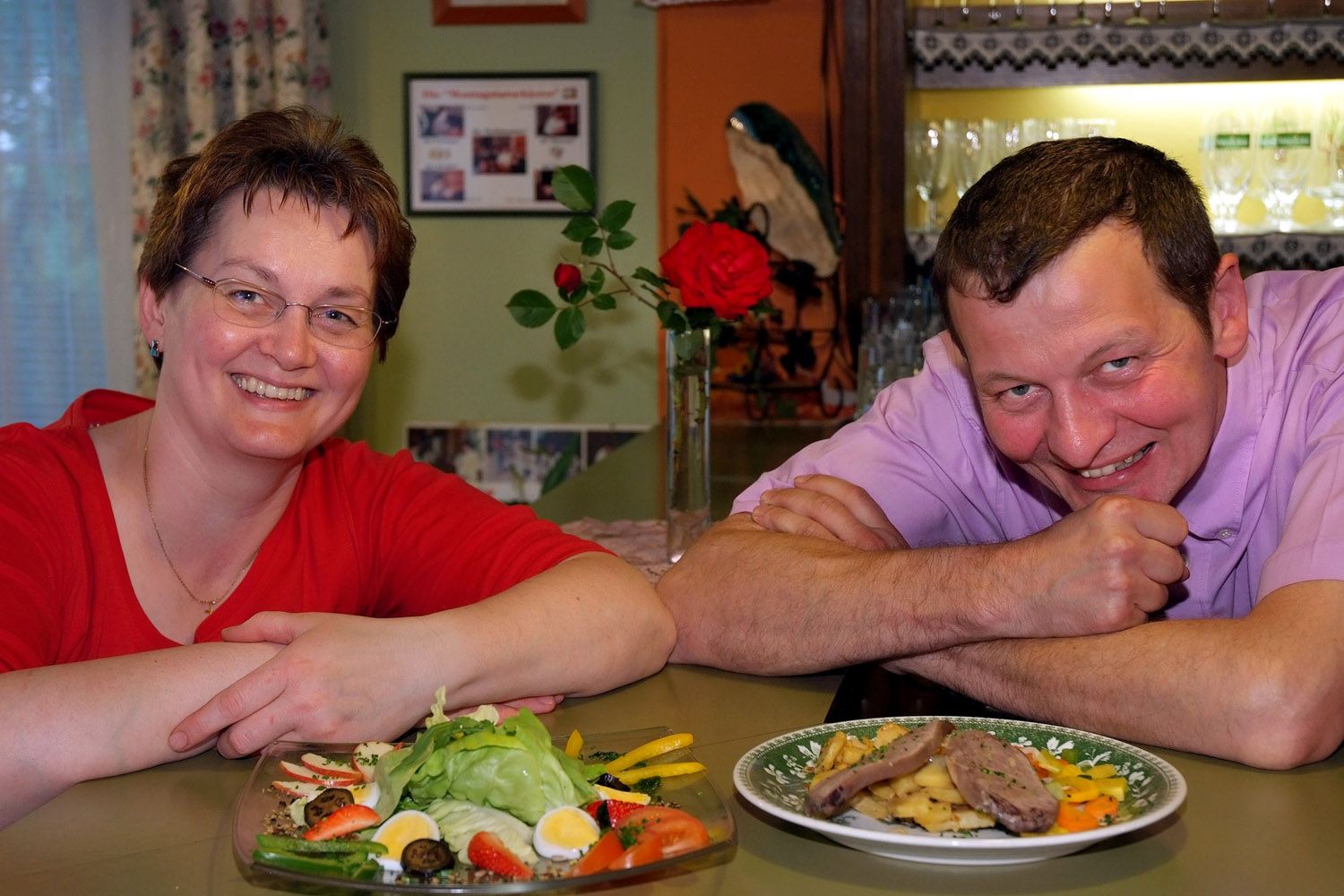 Ein Mann und eine Frau lächeln in die Kamera, vor ihnen stehen Teller mit Essen. Im Hintergrund ist eine rote Rose und ein Regal mit Gläsern zu sehen.