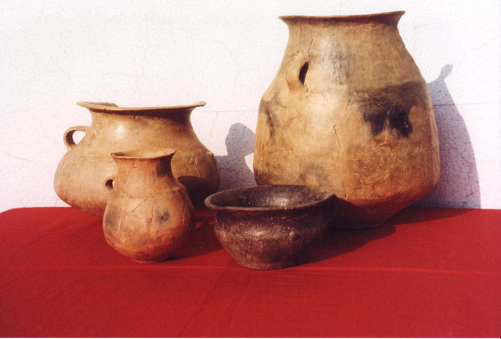 Vier antike Tongefäße auf rotem Tuch vor weißer Wand.