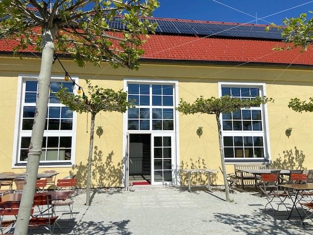 Gelbes Gebäude mit roten Dachziegeln, großen Fenstern und einem Biergarten mit Tischen und Stühlen im Freien.