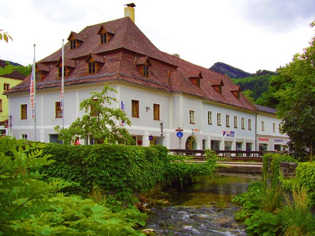 Historisches Gebäude mit rotem Dach an einem Bach, umgeben von grüner Vegetation.