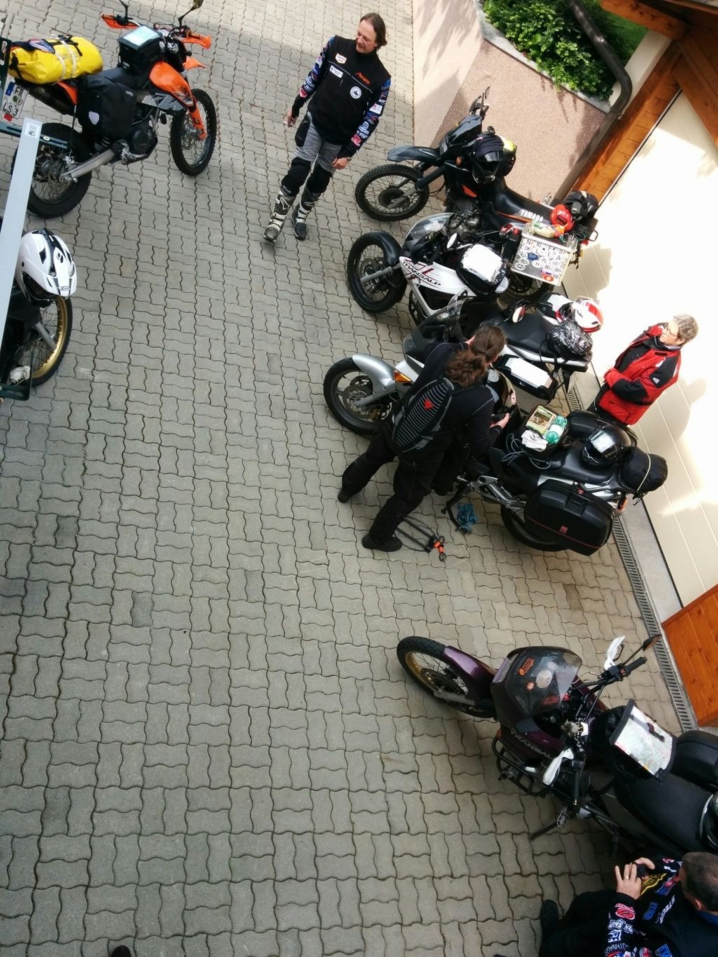 Mehrere Motorräder und Personen auf einem gepflasterten Platz vor einem Gebäude.