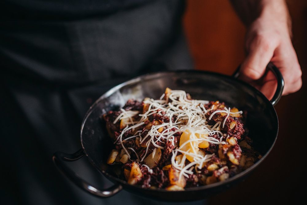 Eine Person hält eine Pfanne mit Blunzengröstl, garniert mit geriebenem Käse.