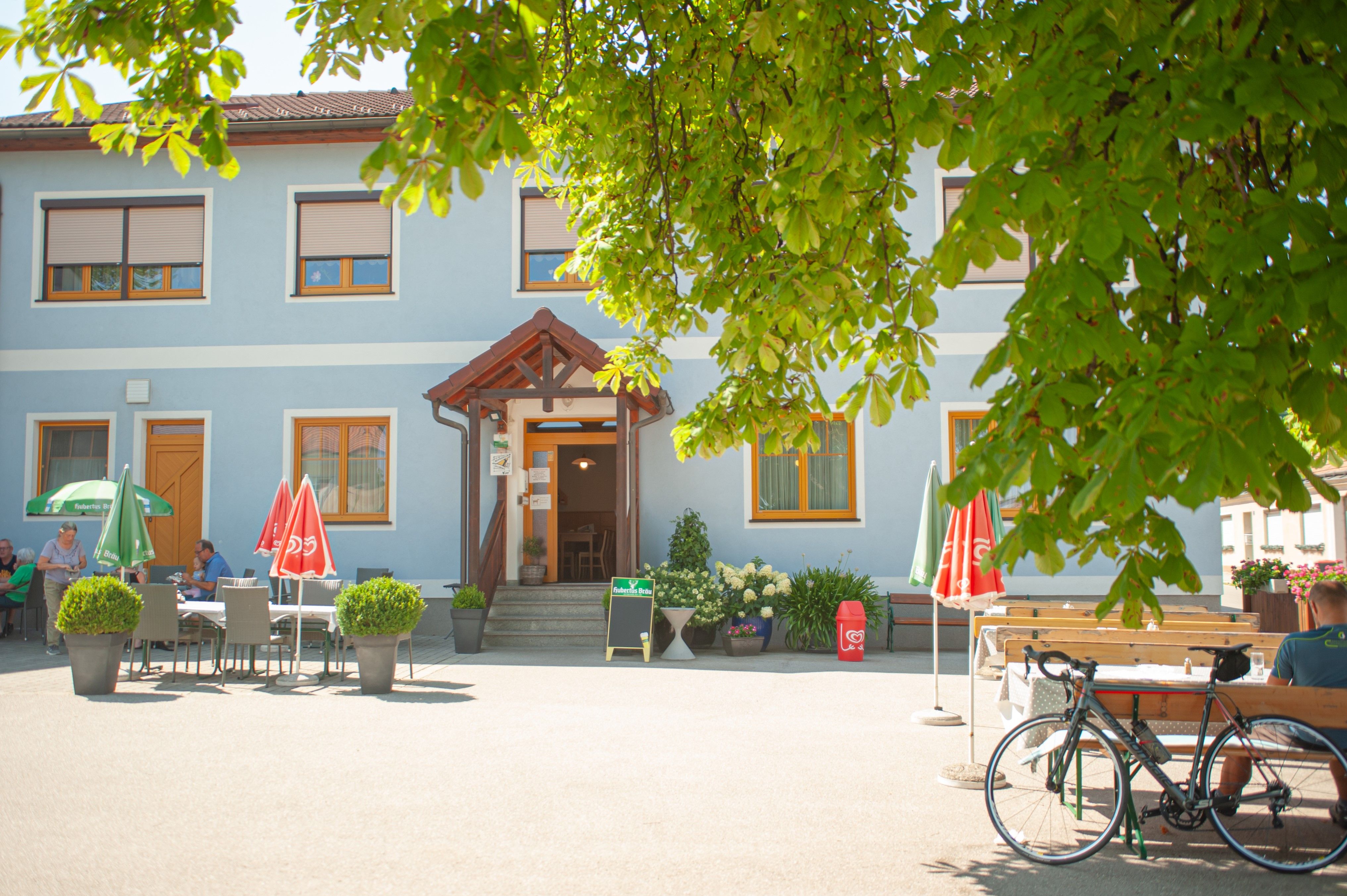 Ein blaues Gasthaus mit Sonnenschirmen und Tischen im Freien, umgeben von grünen Bäumen.