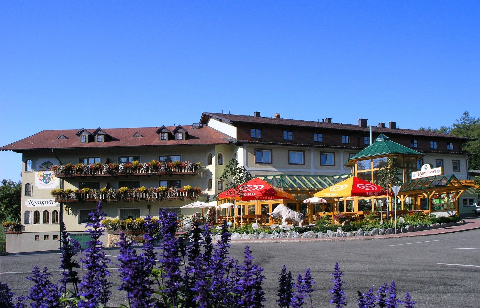 Gasthof Ramswirt mit blühenden Balkonen und Sonnenschirmen vor blauem Himmel.