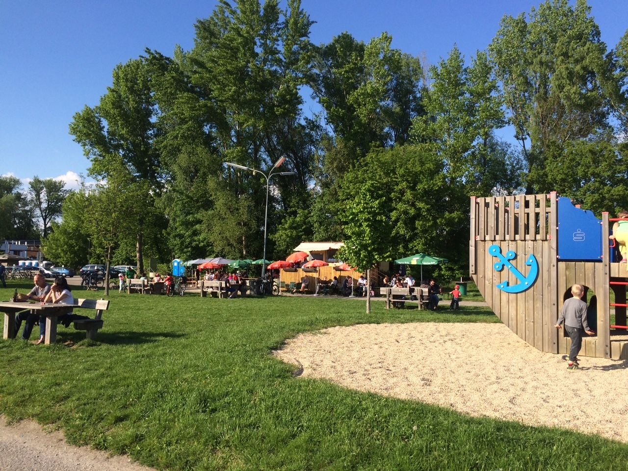 Spielplatz mit Holzklettergerüst und Imbissbereich im Hintergrund, umgeben von Bäumen und Wiese.