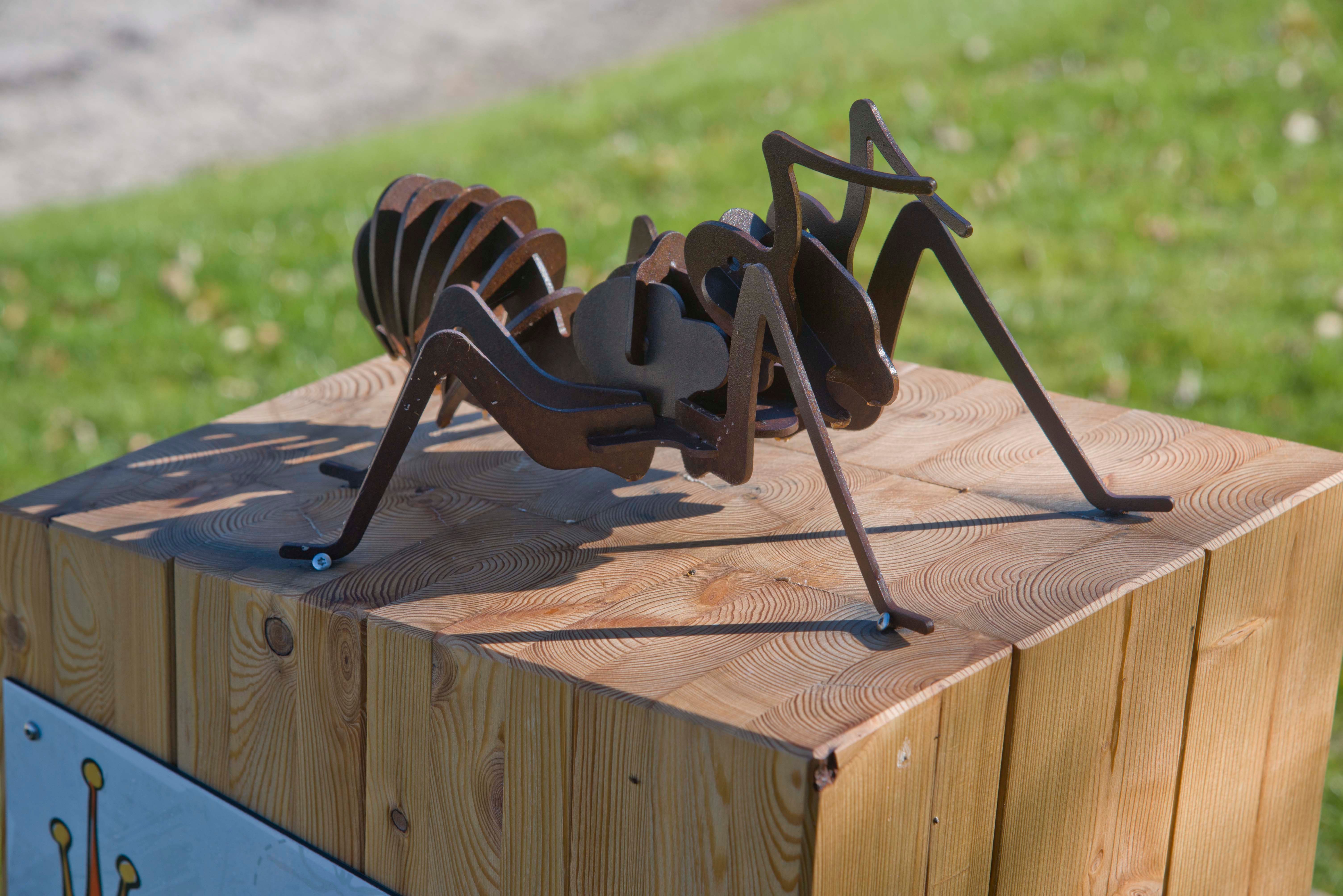 Metallskulptur einer Ameise auf einem Holzblock im Freien.