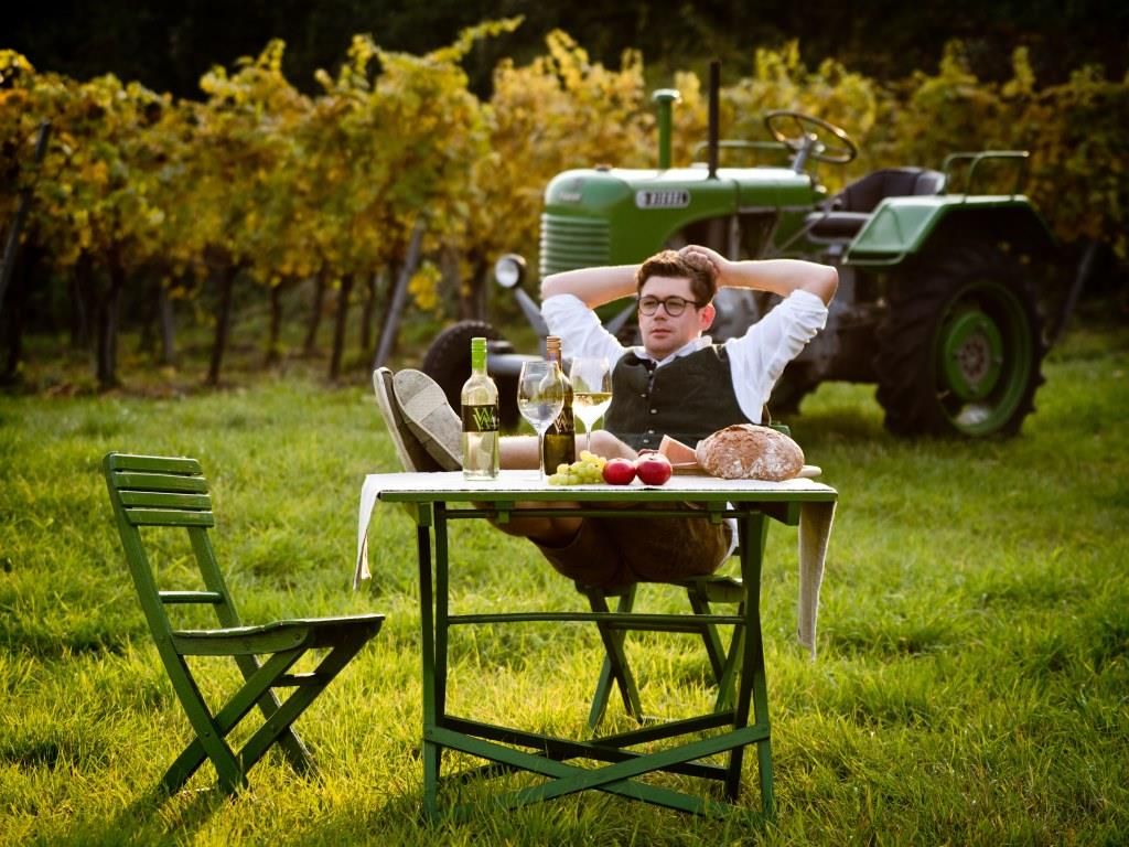 Ein Mann entspannt sich an einem Tisch im Weinberg mit Wein, Brot und Äpfeln. Im Hintergrund steht ein grüner Traktor.