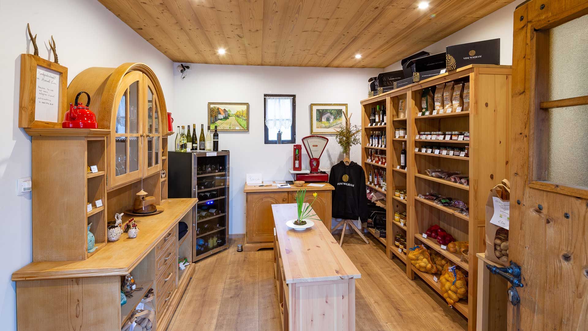 Innenansicht eines kleinen, gemütlichen Ladens mit Holzregalen, gefüllt mit regionalen Produkten wie Wein, Marmelade und Gemüse.