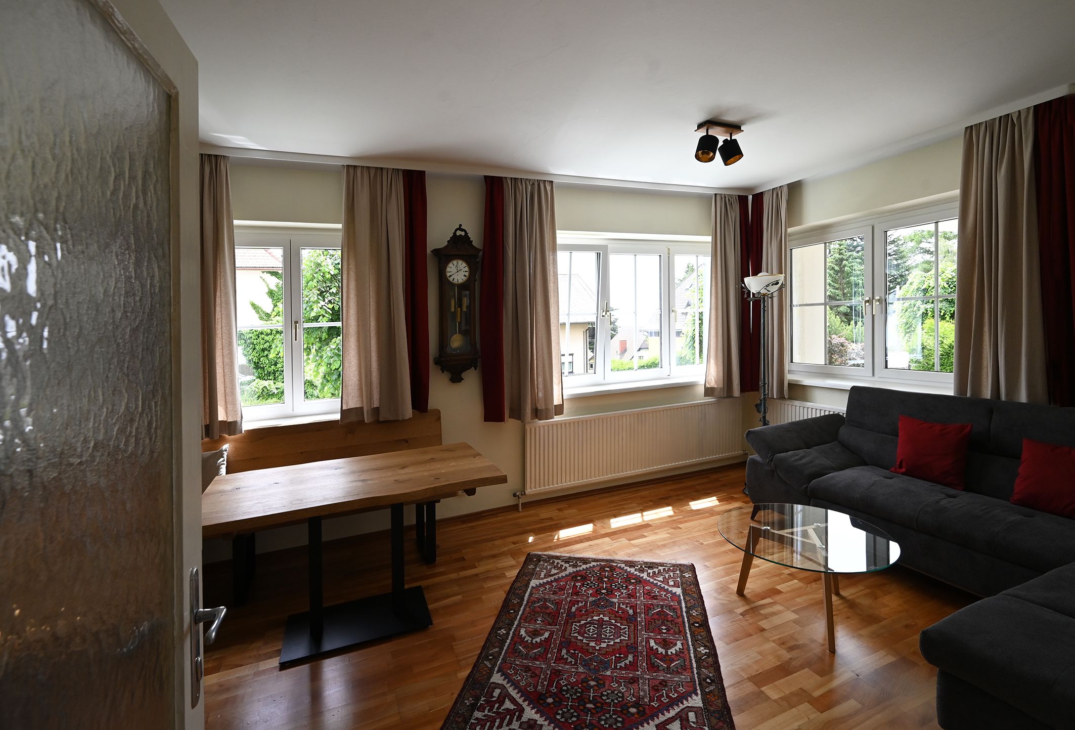 Ein Wohnzimmer mit Holzboden, grauem Sofa, Glastisch, Wanduhr und Teppich. Große Fenster mit Vorhängen lassen viel Licht herein.