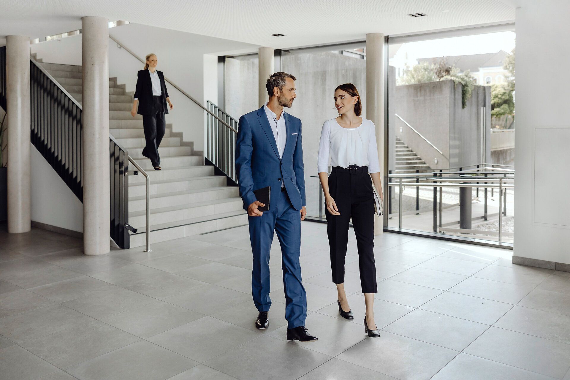 In einem modernen, lichtdurchfluteten Raum schlendern ein Paar in eleganter Kleidung und eine Frau im Hintergrund die Treppe hinunter. Die Atmosphäre ist geprägt von einem Gefühl der Vorfreude und des Austauschs, während die Architektur des Raumes eine harmonische Verbindung von Stil und Funktionalität zeigt.