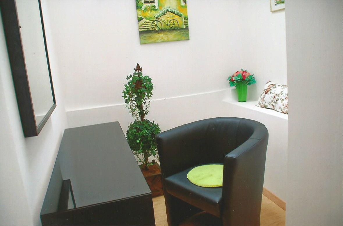 Kleines Gästezimmer mit schwarzem Sessel, grünem Kissen, Spiegel, Pflanzen und Wandbild.