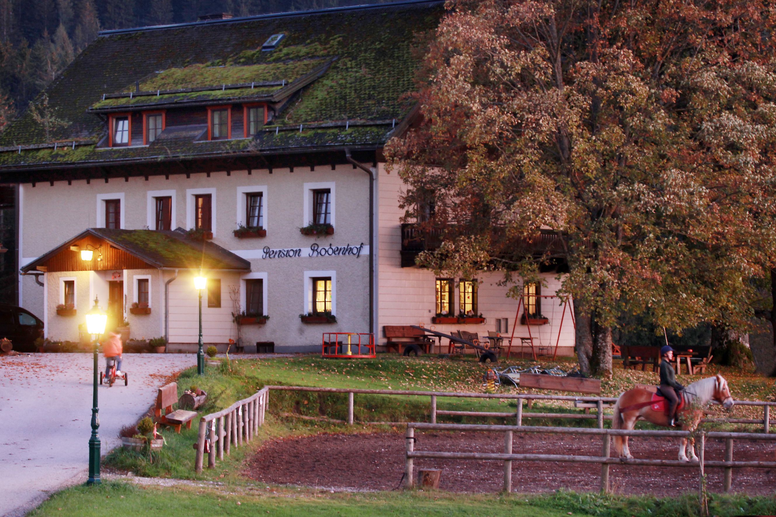 Ein Bauernhof mit beleuchteten Fenstern, einem Reiter auf einem Pferd und einem Kind auf einem Dreirad im Vordergrund.