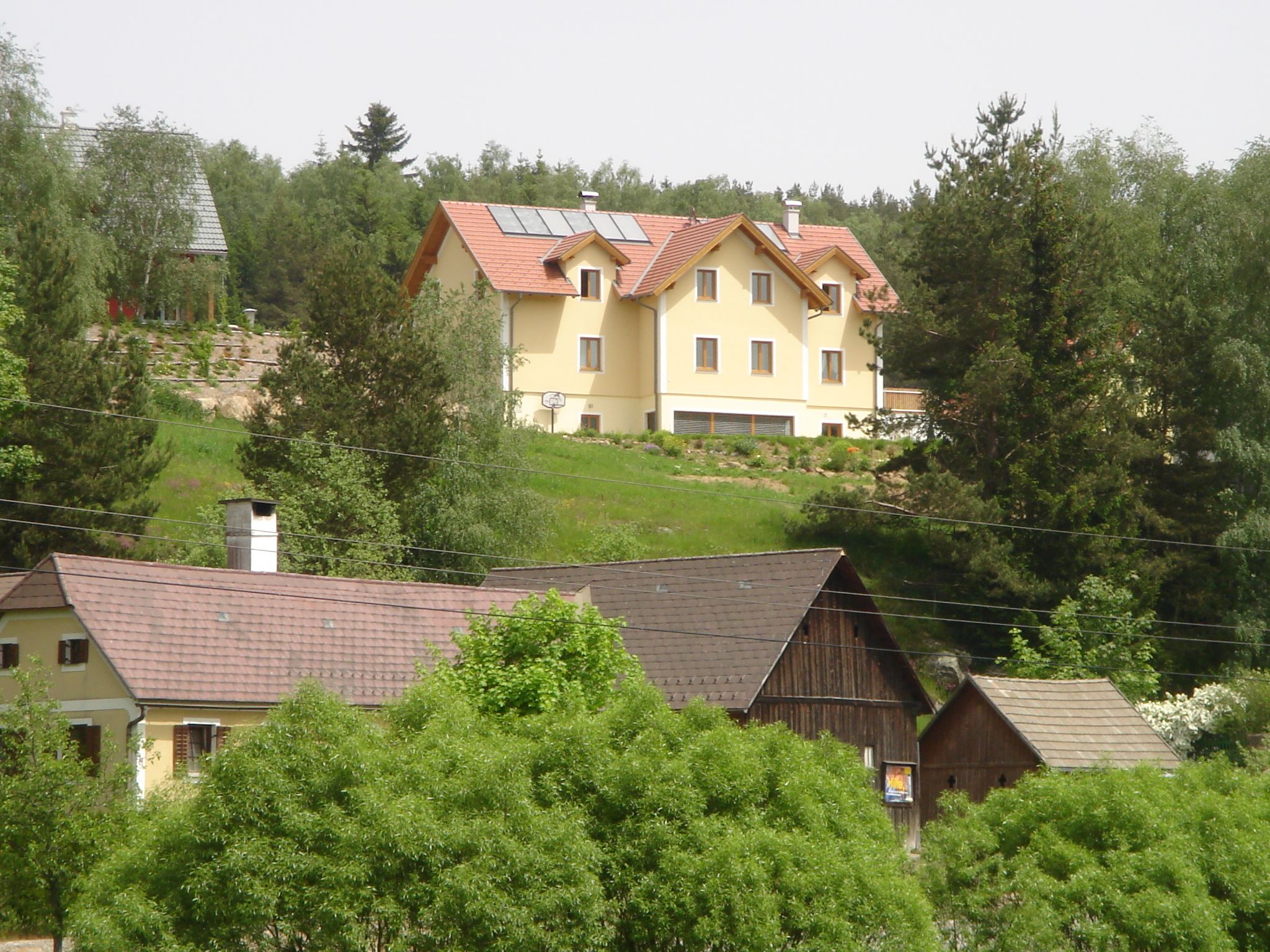 Ein großes gelbes Haus mit Solarpanelen auf dem Dach steht auf einem Hügel, umgeben von Bäumen und kleineren Gebäuden im Vordergrund.