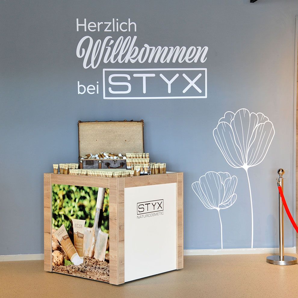 Ein Display mit STYX Naturkosmetik-Produkten vor einer grauen Wand mit Blumenzeichnung und Willkommensgruß.